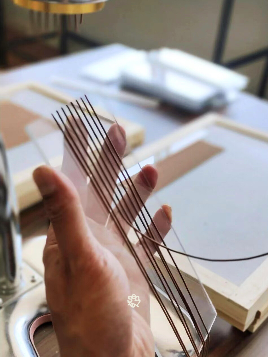 Close-up of a person's hand holding a piece of thin, curved glass or plastic with embedded thin black lines, with a workshop or crafting area in the background.