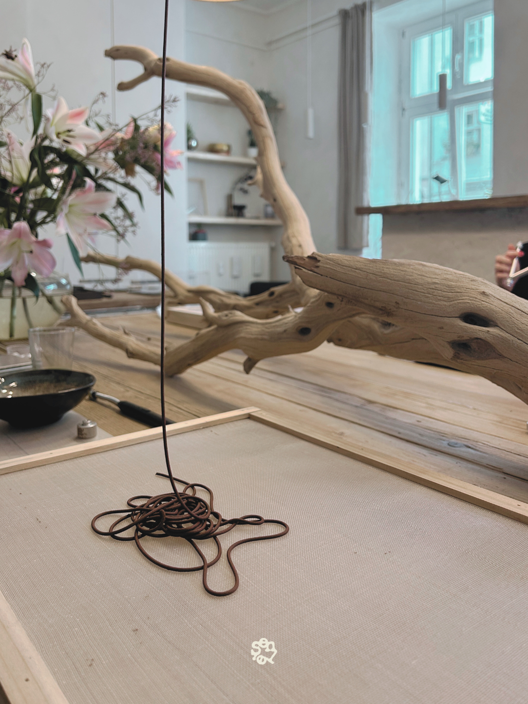 A table decorated with a large piece of driftwood, pink and white flowers in a vase, a black bowl, and various tools. A mirror reflects the room, showing a window with curtains and shelves with small decorative items.