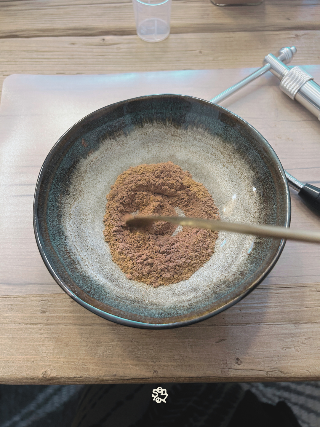 A black bowl containing a mound of brown powder, placed on a light wooden surface, with a metal tool and a clear plastic container nearby.