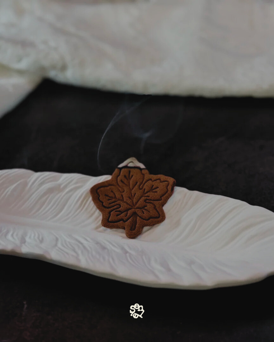 A brown maple leaf-shaped incense cone on a white, textured ceramic dish with smoke rising from it.