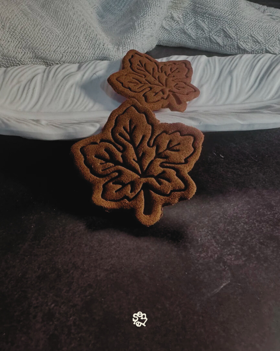 Two brown maple leaf-shaped cookies with leaf vein patterns, placed on a dark surface in front of a white textured pillow.