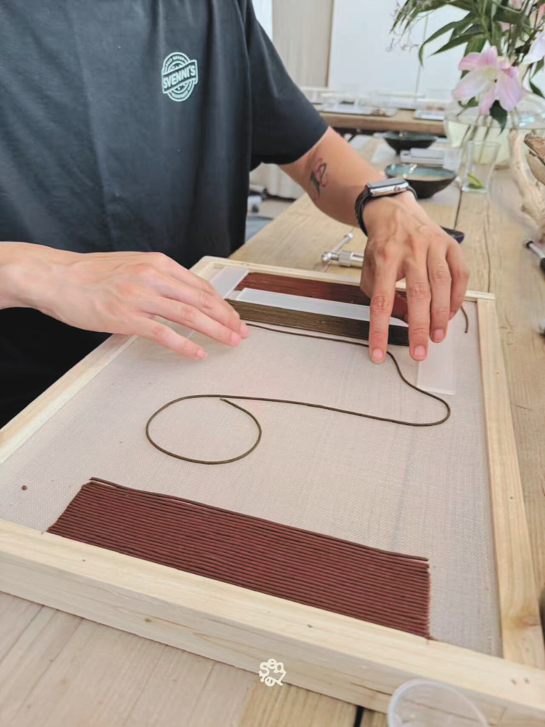 Person assembling a lamp or decorative piece with strips of brown and red material on a wooden frame, using a thin cord, in a workspace with flowers and tools in the background.