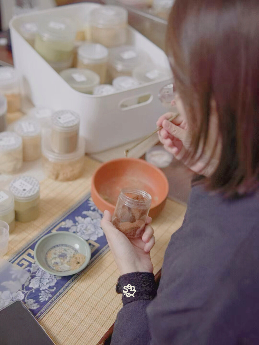 Person handling small jars of powder or spice, using a small scoop or brush over a table with more jars, bowls, and a tray with floral design.