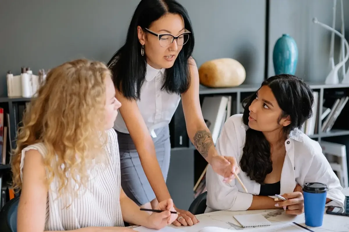Person standing behind two colleagues sitting at a table and reviewing documents on the table.