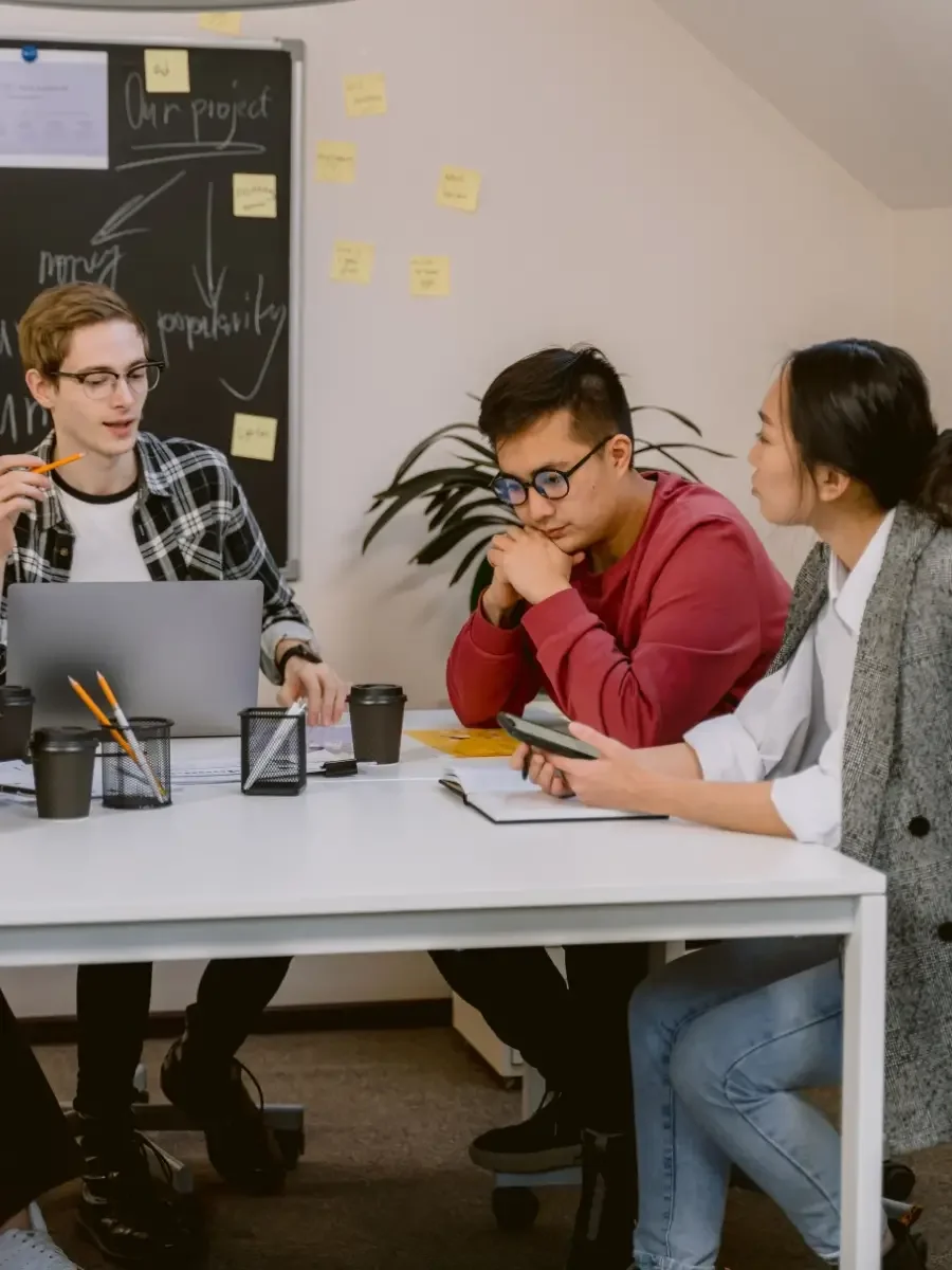 Three people sit around a table having a discussion in an office with a chalkboard covered in sticky notes.