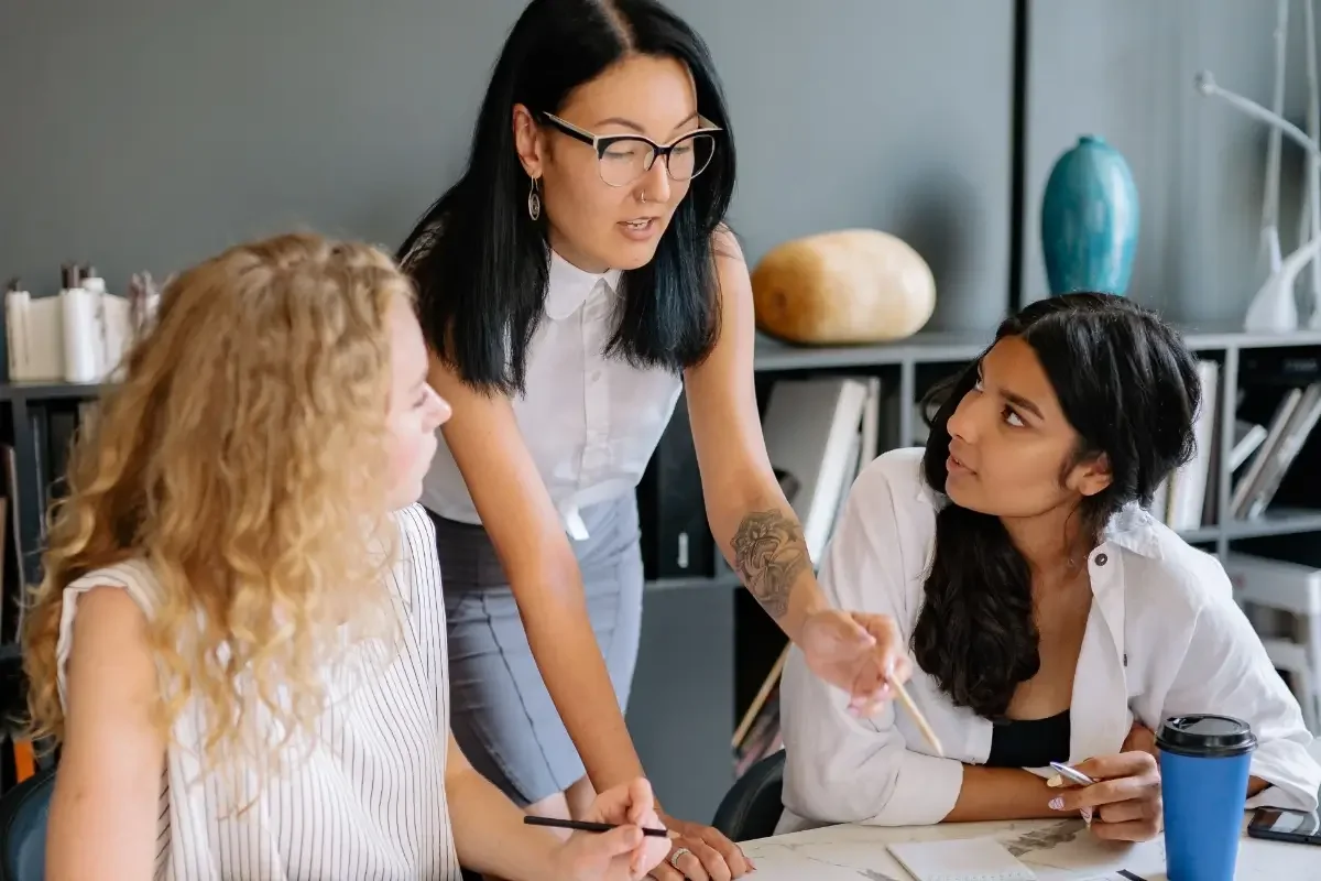 Person standing behind two colleagues sitting at a table and reviewing documents on the table.