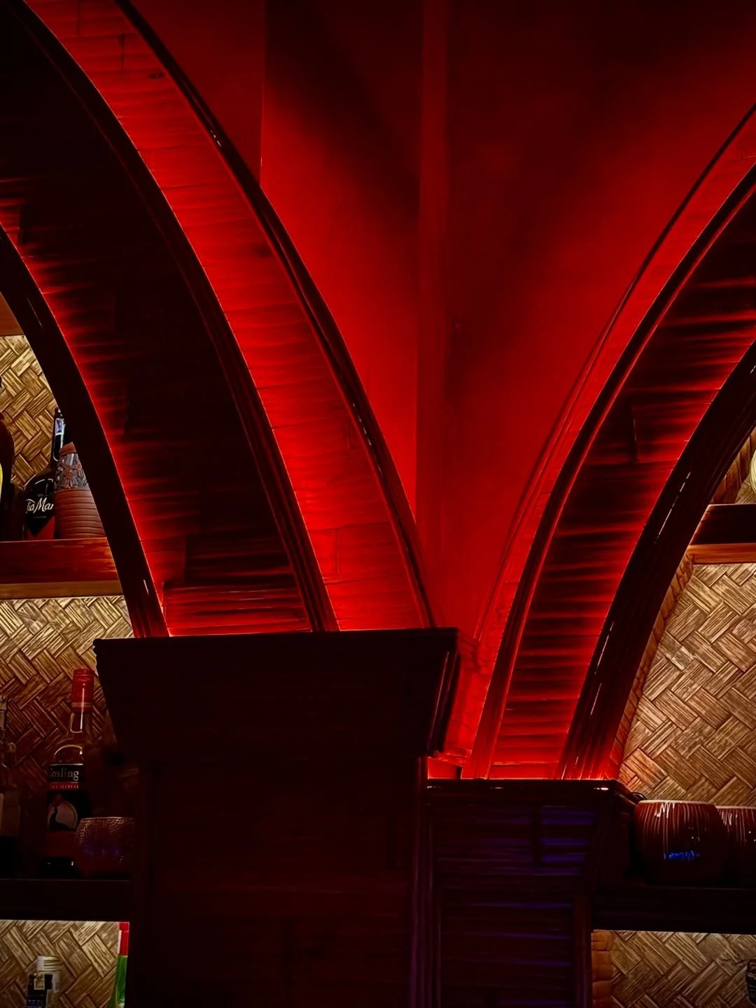 Interior of a structure with curved wooden beams illuminated by red lighting, and a wall with woven bamboo decor.
