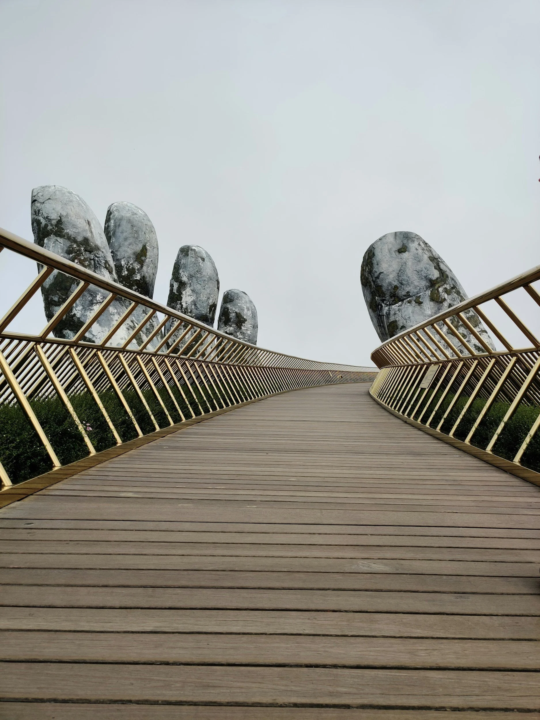 Wooden walkway with golden railings leading towards large, moss-covered rocks under a cloudy sky.