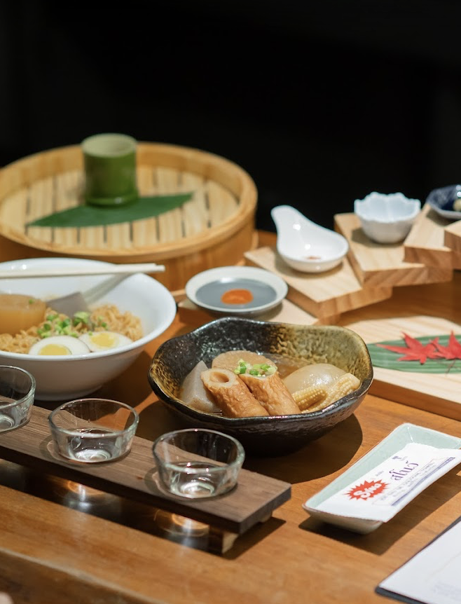 Japanese meal with noodles, fish cake, tofu, and scallions on a wooden table, with small dipping bowls and a bamboo steamer in the background.