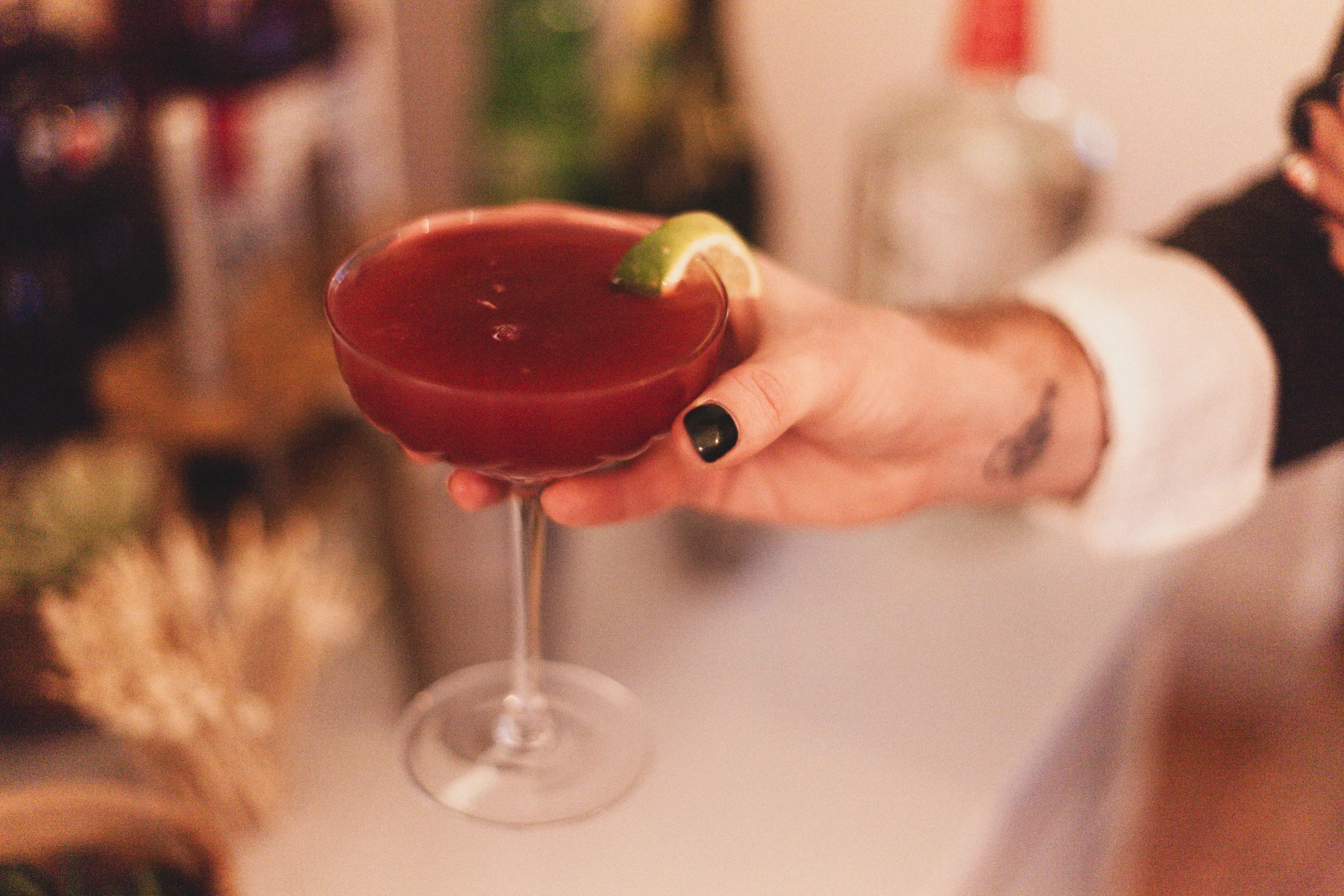 A person holding a red cocktail in a stemmed glass with a lime wedge garnish