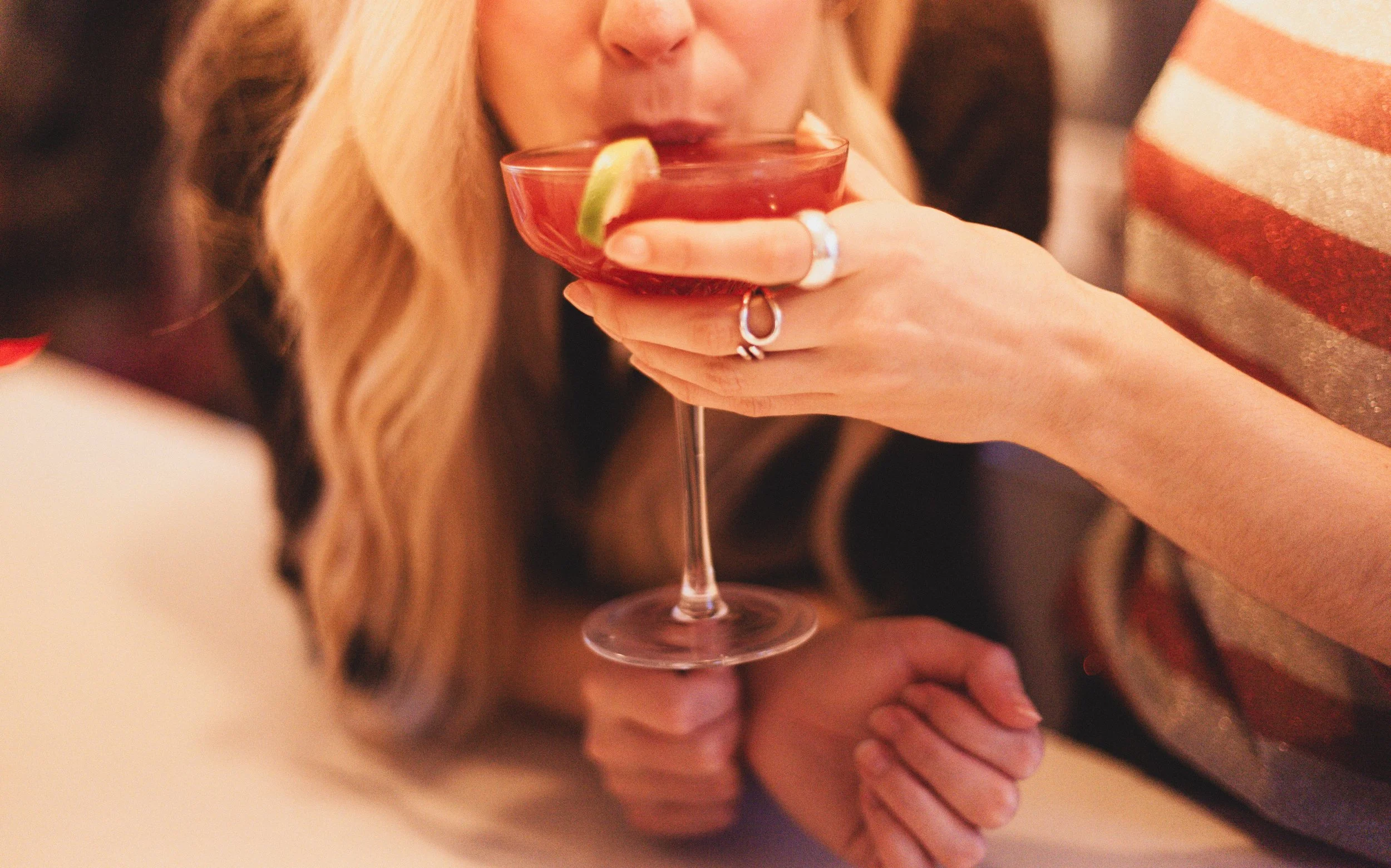 Close-up of a woman with blonde hair drinking a red cocktail with a lime wedge garnish at a social gathering.