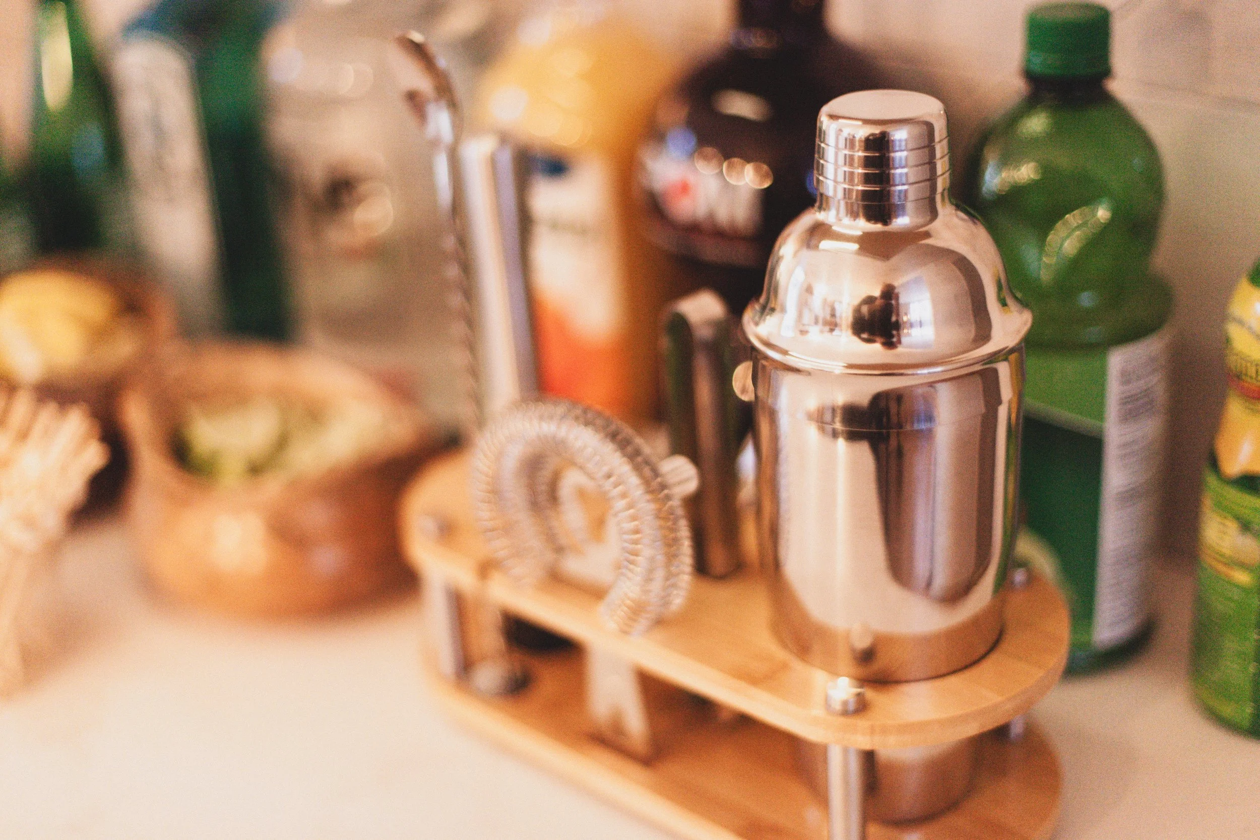 A vintage-style cocktail shaker on a wooden stand, surrounded by various bottles and snacks on a counter.