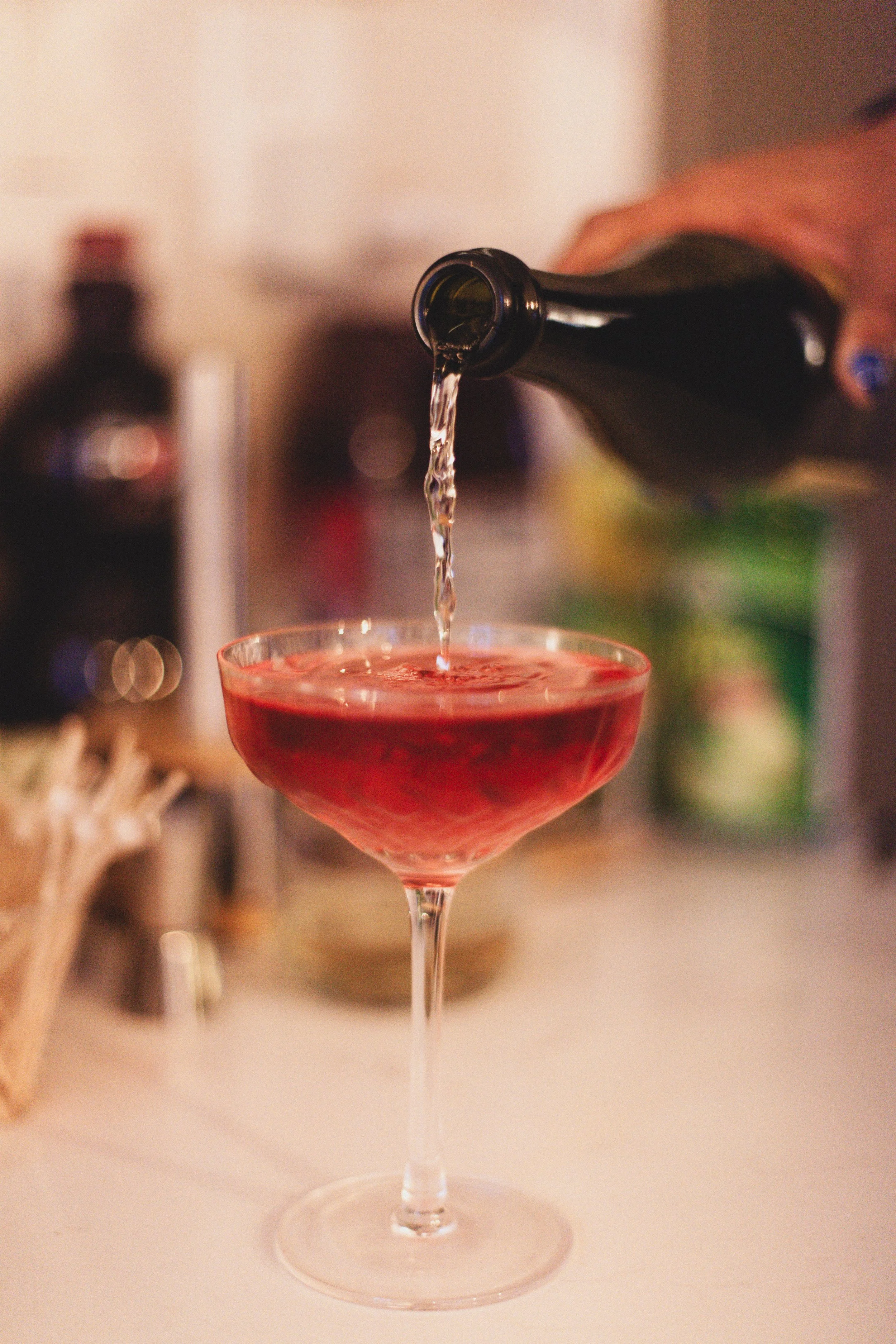 A glass of red cocktail with clear liquid being poured from a bottle.