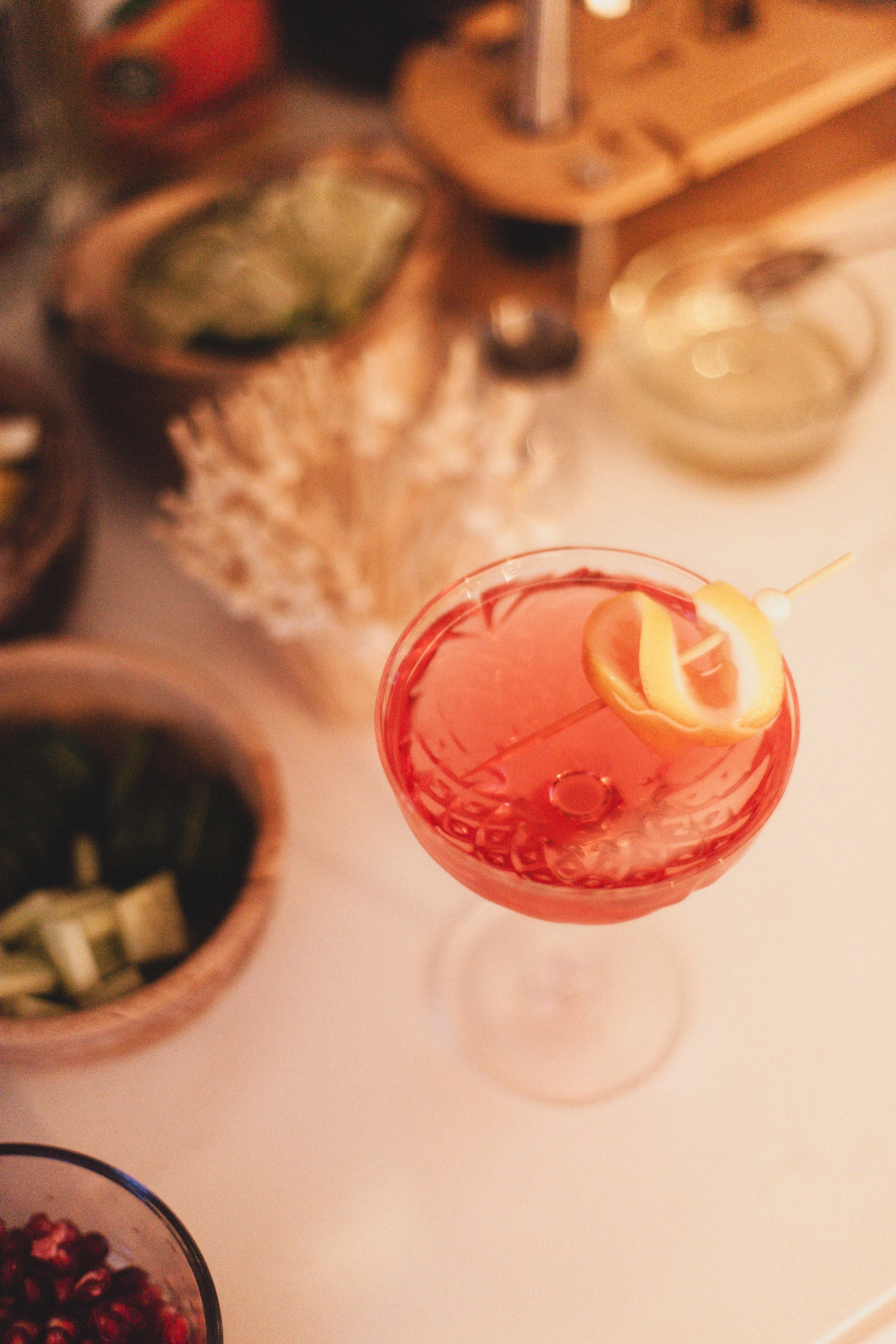 A pink cocktail with a lemon twist garnish on a stick, placed on a white table.