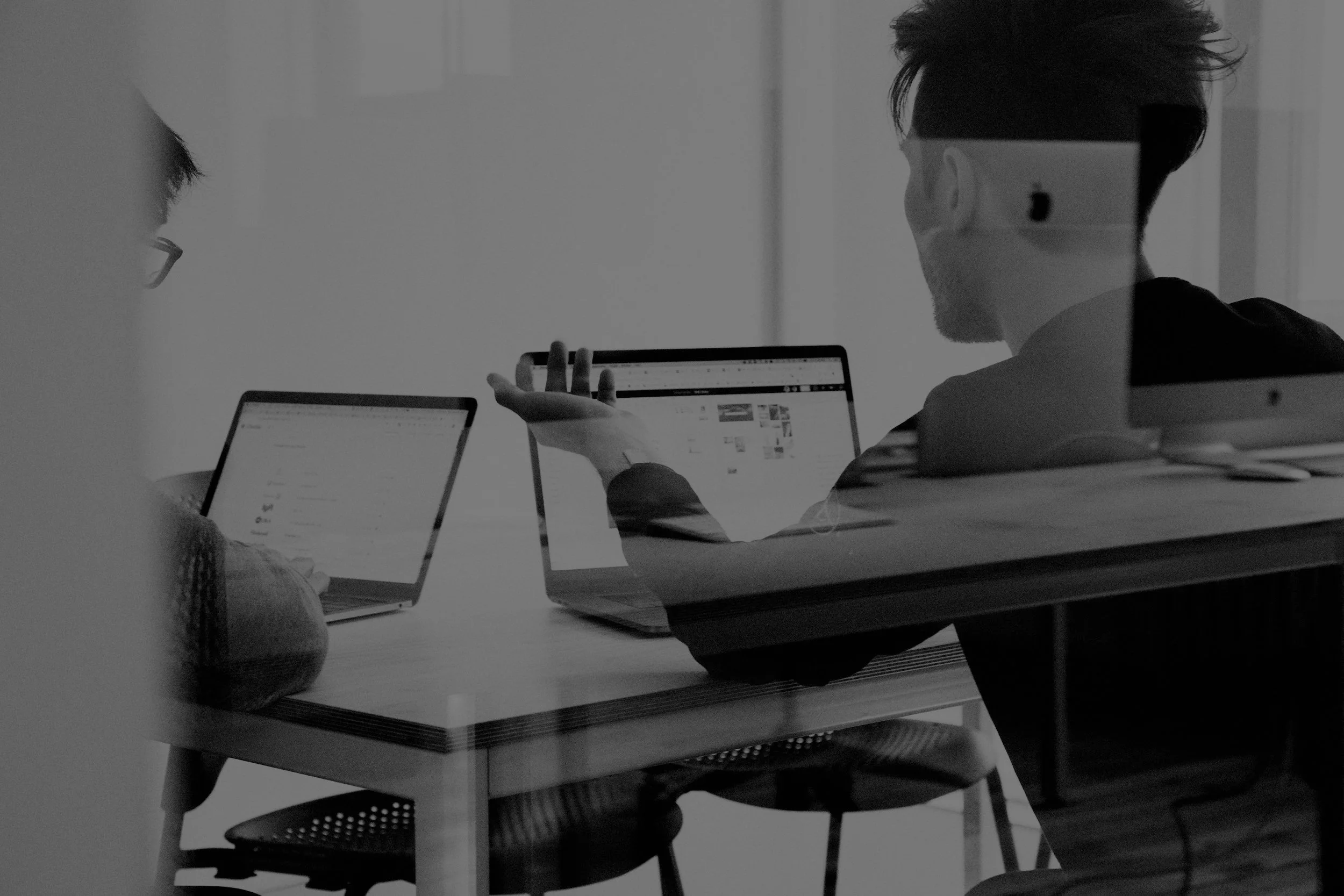 Two people are sitting at a table with laptops, engaged in conversation or discussion in an office setting.