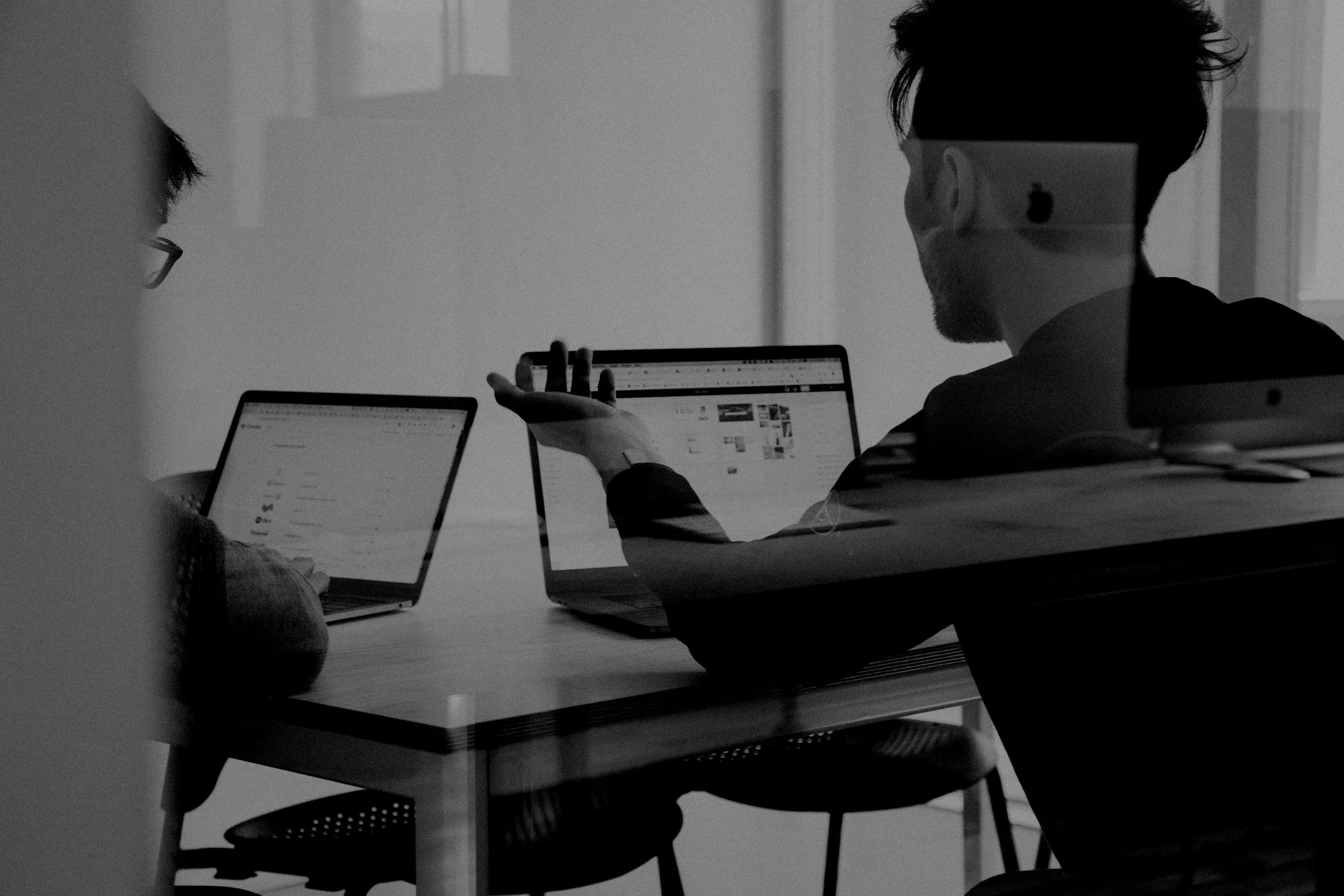 Two people sitting at a table in a meeting room, using laptops, with one person gesturing to the other's screen.