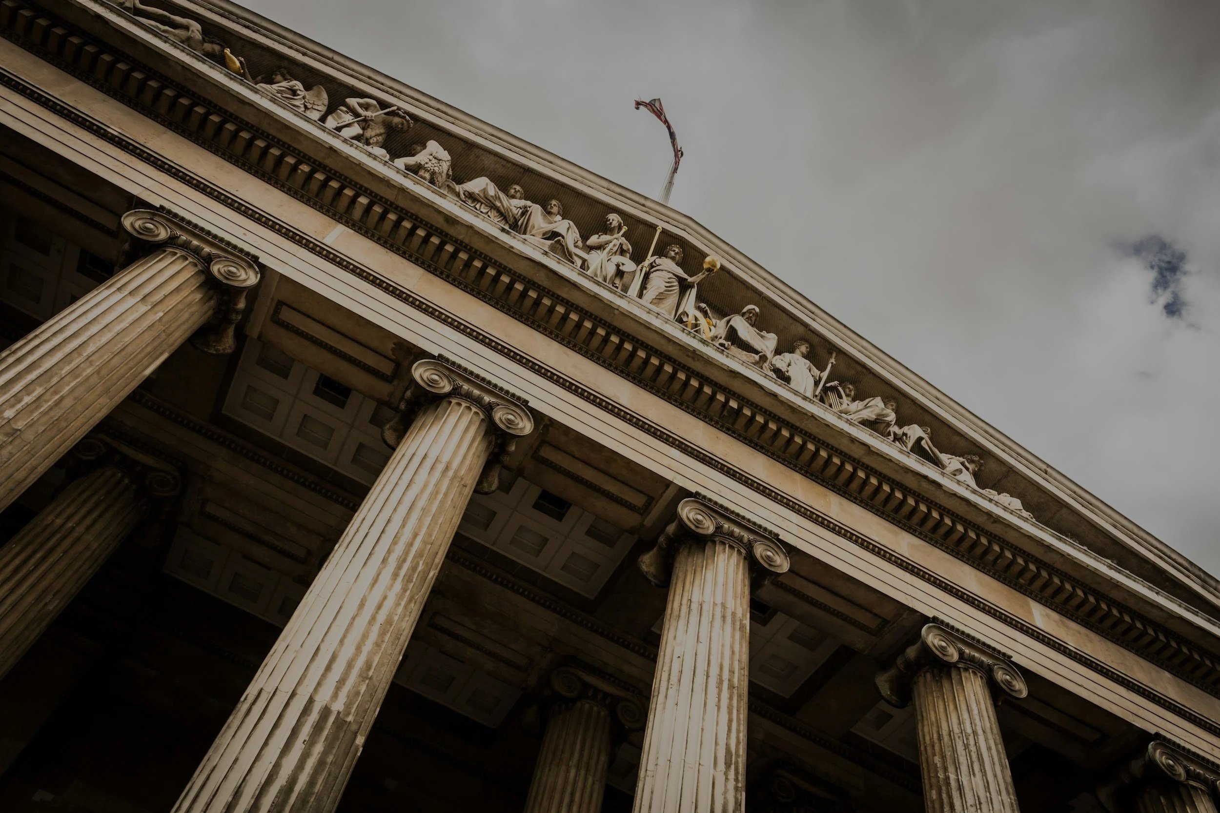 Low-angle view of the front of a classical government building with tall, fluted columns and detailed sculptures on the architrave, against a cloudy sky.