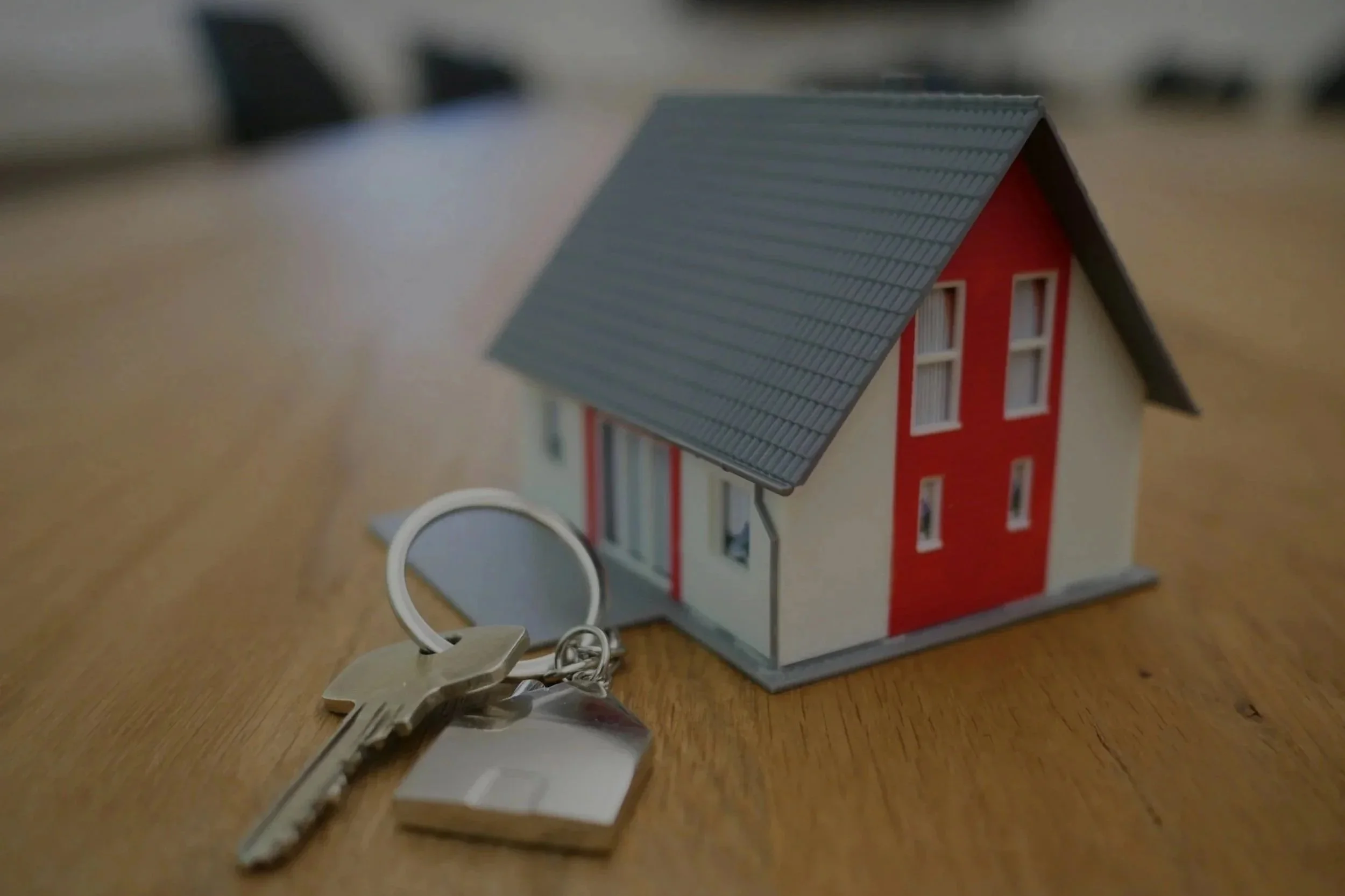 A small model house with a gray roof and red and white walls, placed on a wooden surface, with a set of keys in the foreground.