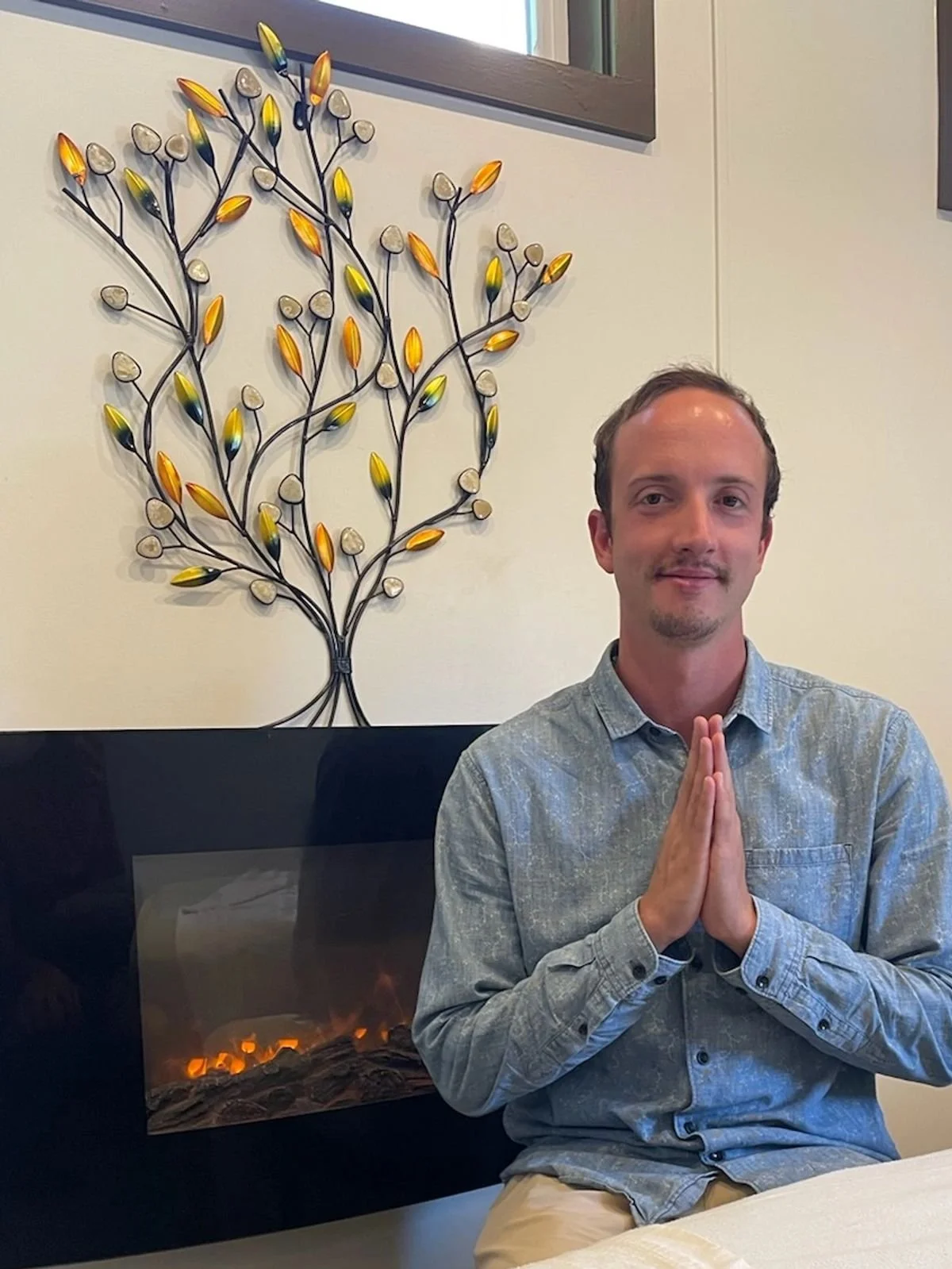 A man sitting with hands pressed together in a prayer-like gesture, in front of a lit fireplace and a decorative wall piece resembling a tree with colorful leaves.