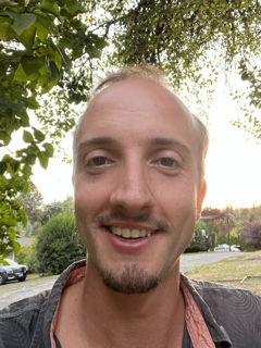 A smiling man with short blonde hair, light skin, and a goatee, outdoors with trees and cars in the background.