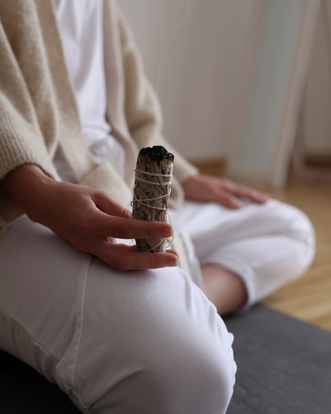 Person sitting cross-legged on a mat holds a smudging bundle, with a blurred background of a light room.