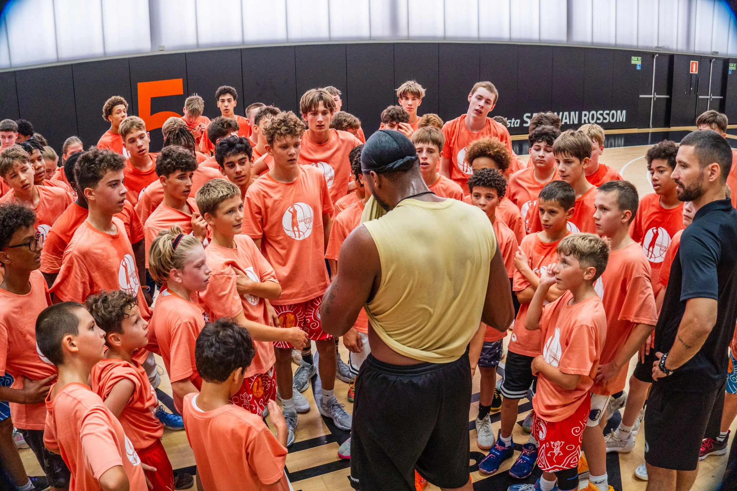 A group of young basketball players wearing orange shirts listening to coach in a gym.