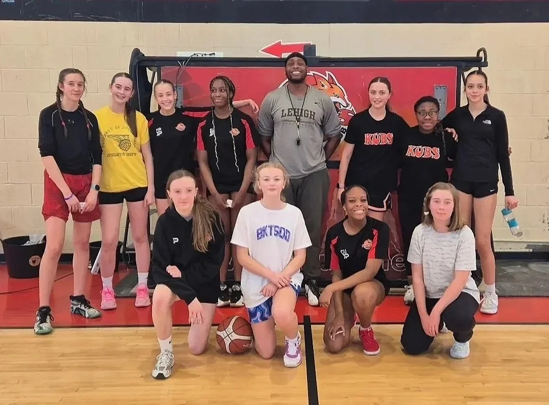 A girls' basketball team posing for a photo in a gymnasium. The team is standing and kneeling in front of a basketball hoop, with a coach in the center. The players are wearing athletic clothing with some in black, red, and yellow uniforms, and they 