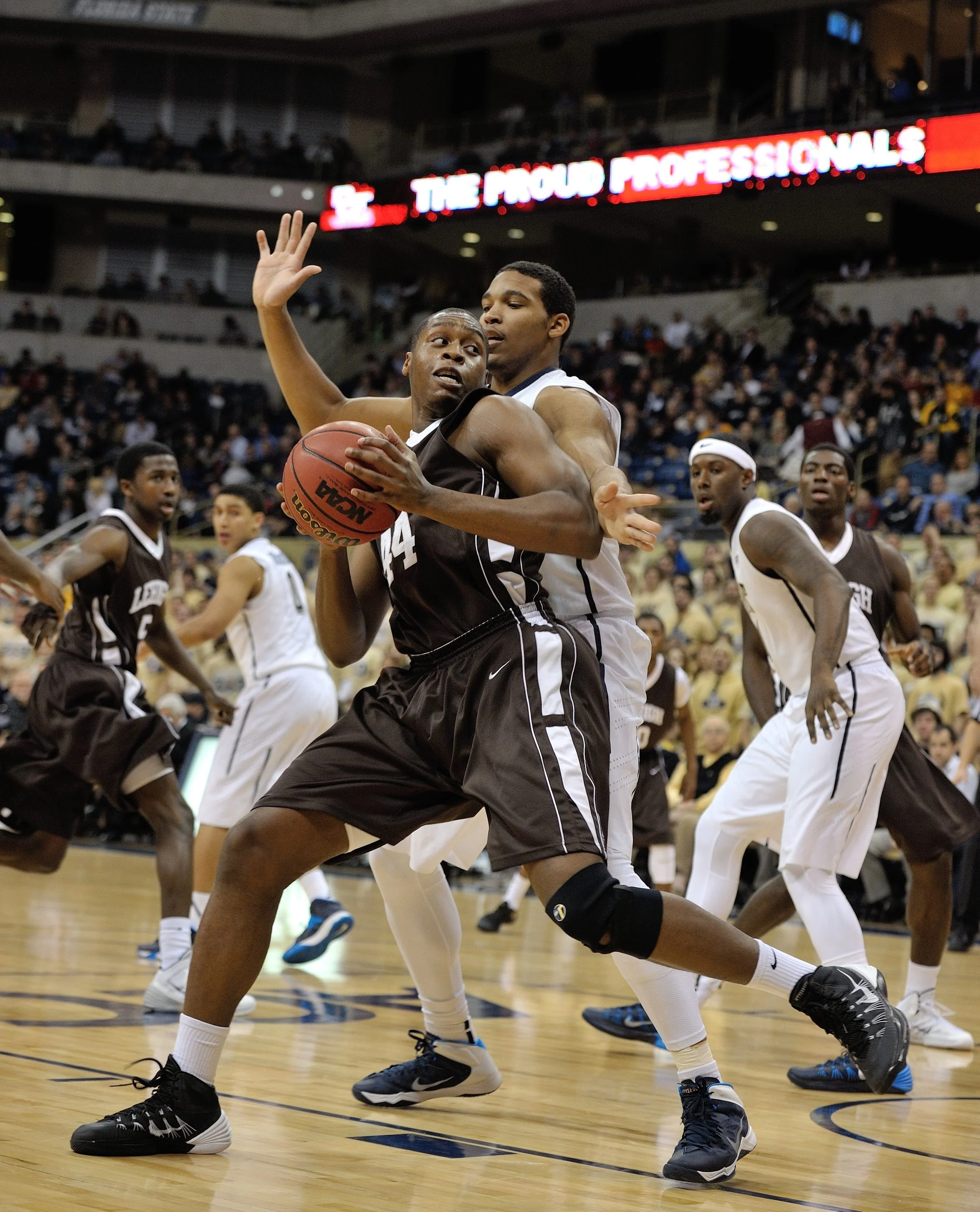 Basketball game with players battling for possession of the ball, one player in black holding the ball while a player in white defends him, in an indoor arena filled with spectators.