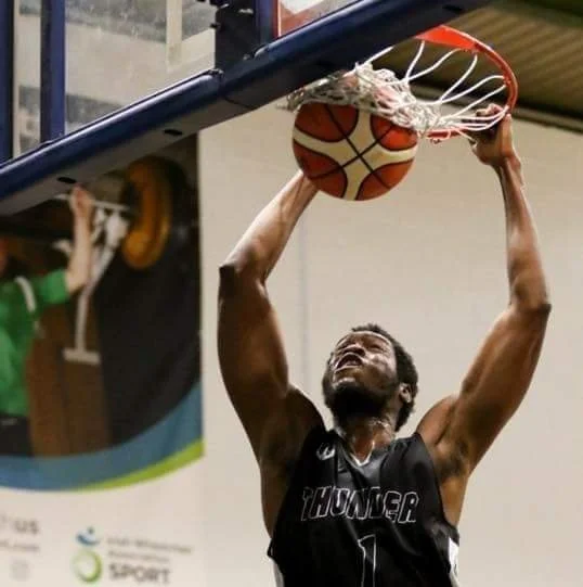 A basketball player in a black 'Thunder' jersey dunking a basketball through the hoop during a game in an indoor gym.