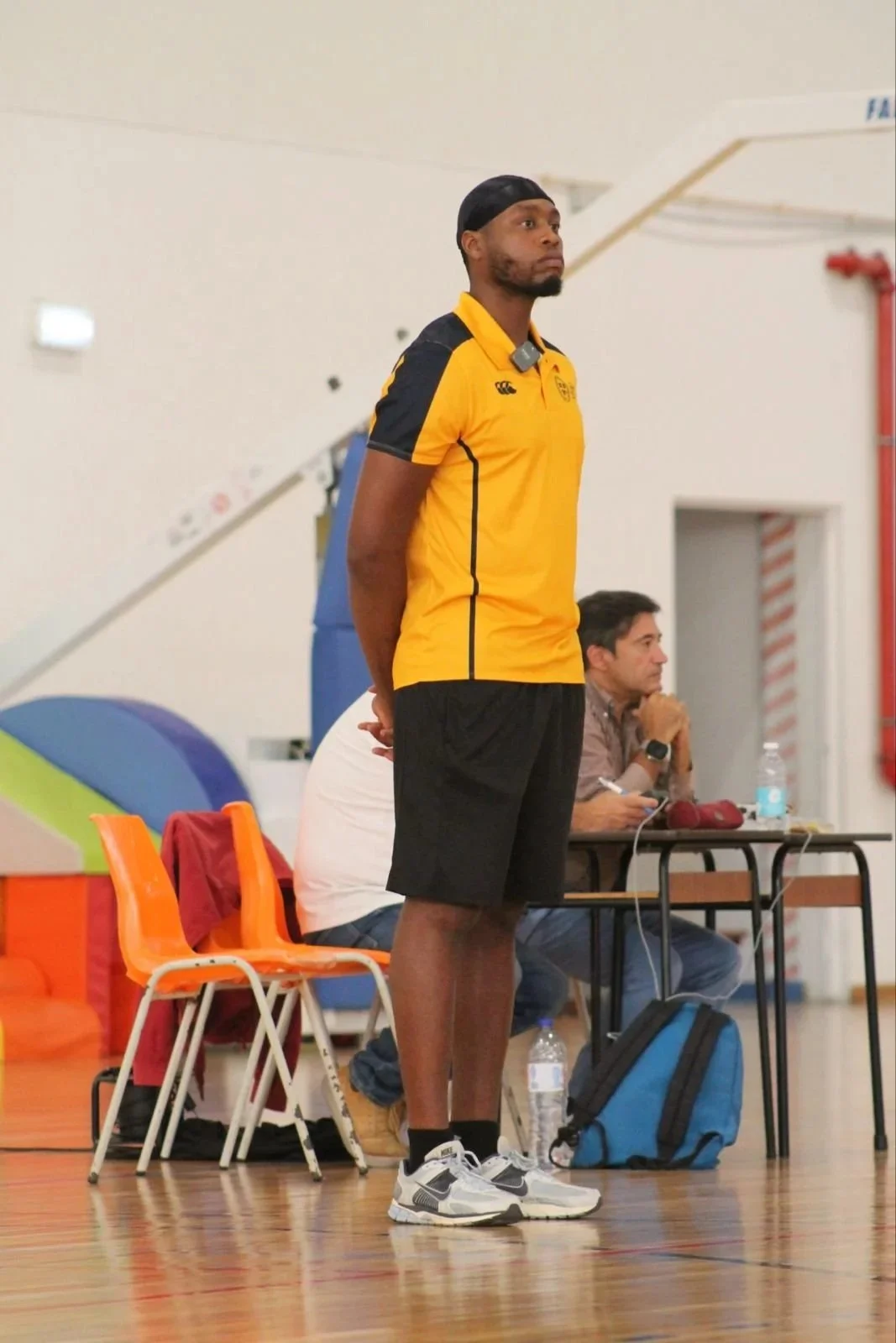 A man in a yellow and black sports shirt, black shorts, and sneakers standing with hands behind his back in an indoor gym. Behind him, a person seated at a table with water bottles, a notebook, and electronic devices is visible.