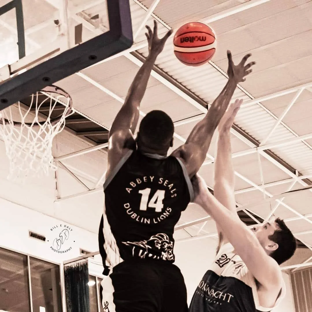 A basketball player in a black jersey with the number 14 is jumping to make a shot at the basket, while a defender in a white jersey attempts to block the shot. Credit: Bill E Daly Sport
