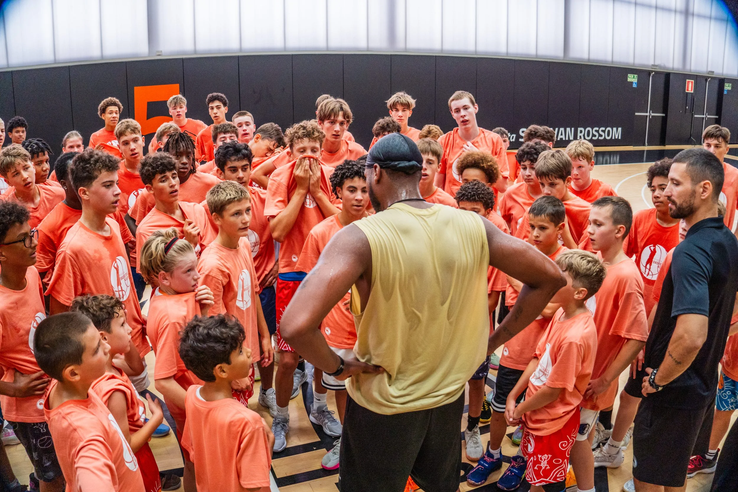 Basketball coach giving instructions to a large group of young players in an indoor gym.