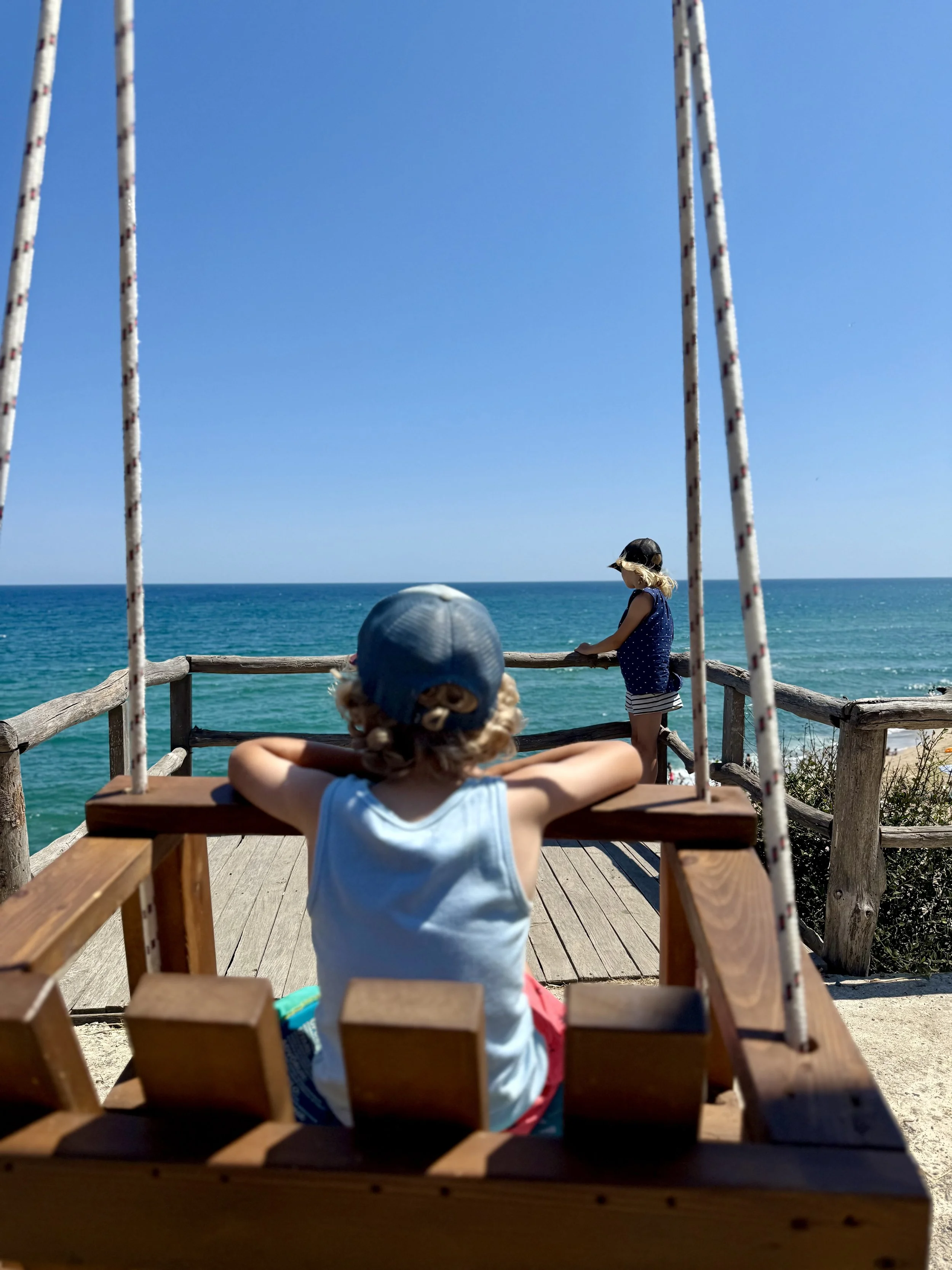 Zwei Kinder auf einer Holzschaukel am Strand mit Blick aufs Meer, eines sitzt auf der Schaukel und schaut aufs Wasser, das andere steht auf der Plattform und schaut in die Ferne.