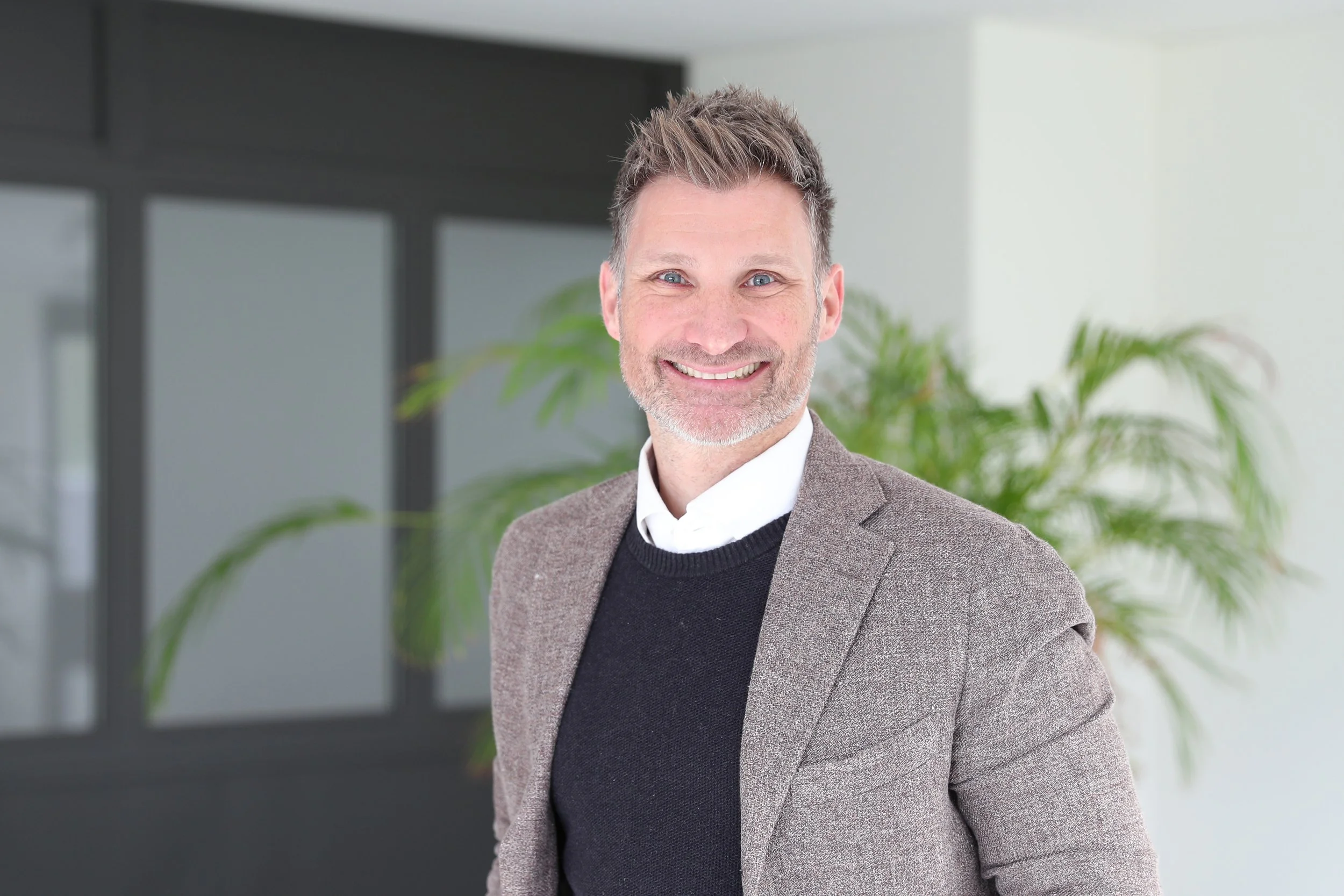 Ein lachender Mann in einem modernen Büro, trägt einen Grauen Anzug mit schwarzem Pullover und weißem Hemd. Im Hintergrund sind large Fenster und eine grüne Pflanze sichtbar.