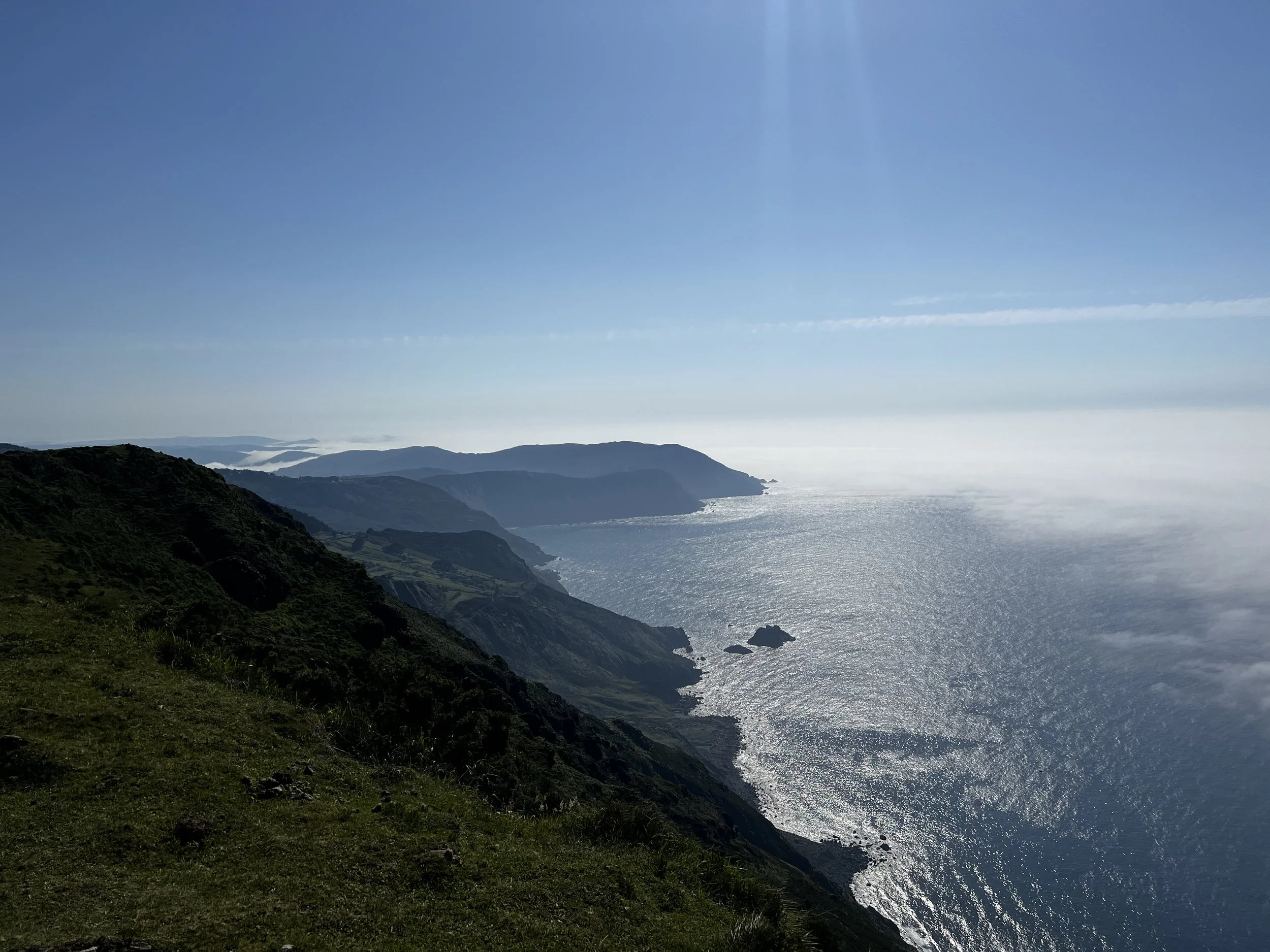 Küstenlandschaft mit grünen Klippen, Blick auf das Meer und den Himmel, Sonnenlicht spiegelnd auf der Wasseroberfläche, am Tag helle Himmel, leichter Nebel im Hintergrund.