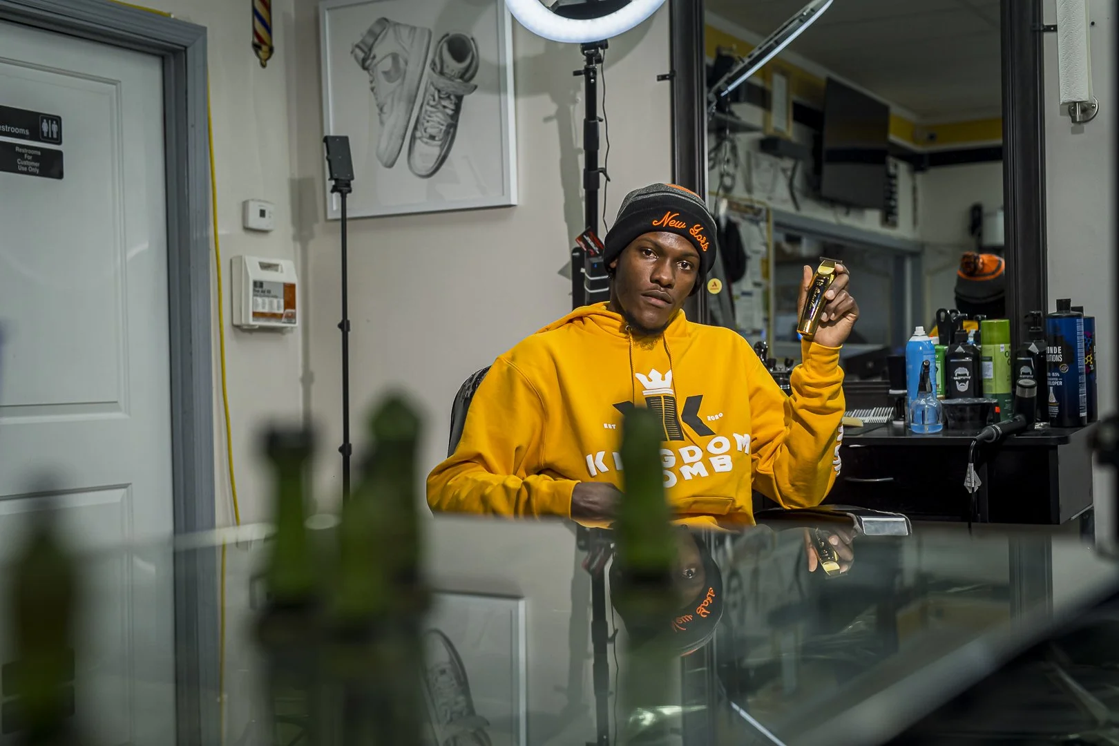 A young man with dark skin wearing a black beanie and yellow hoodie with white and gold lettering sitting at a barber's or salon's workspace, holding a hair clipper. The background shows shelves with grooming products, a mirror, and studio lighting e