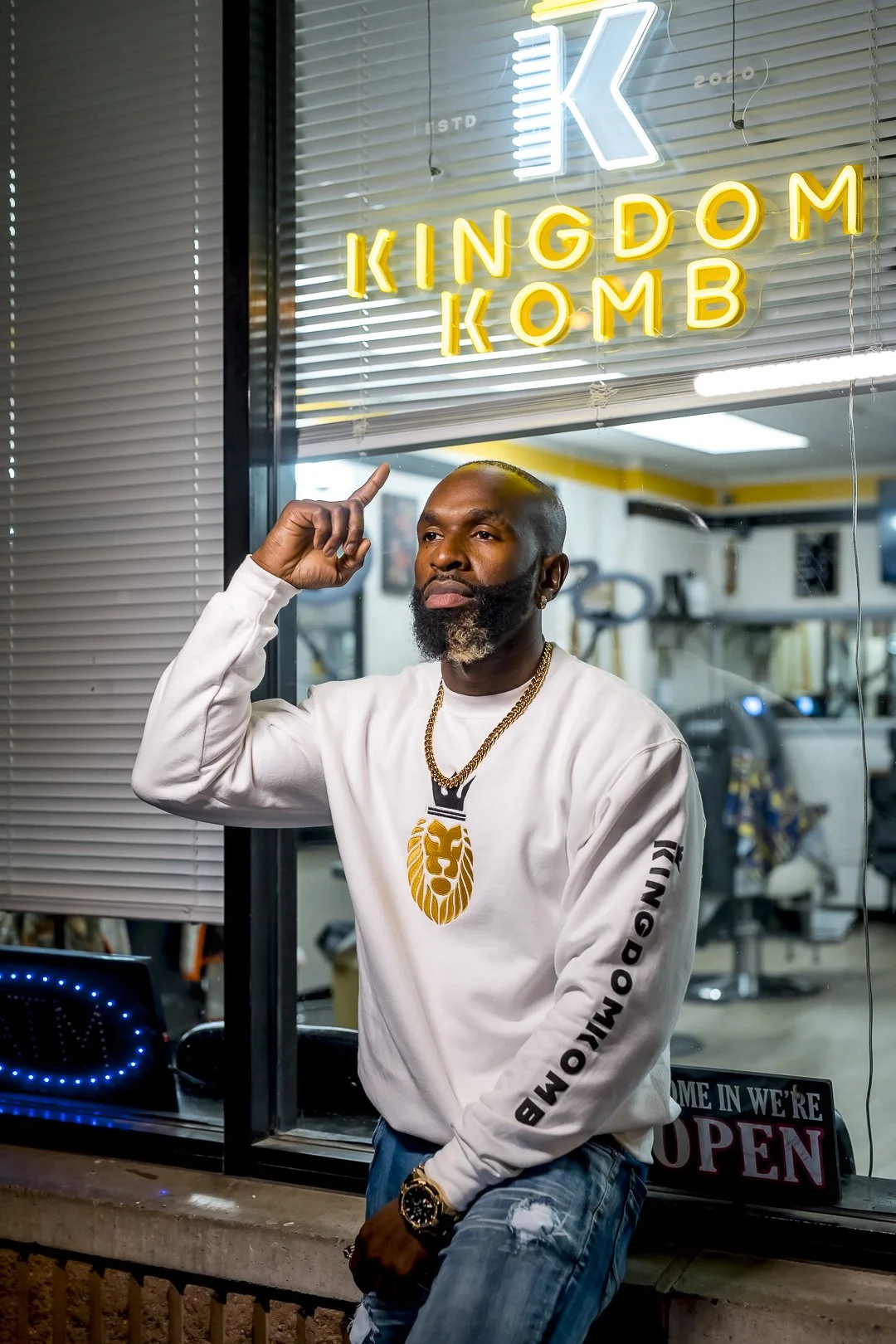 A man standing outside a barber shop named 'Kingdom Komb' at night, pointing up at the shop's neon sign, wearing a white sweatshirt with gold lion and text, ripped jeans, and a gold chain.