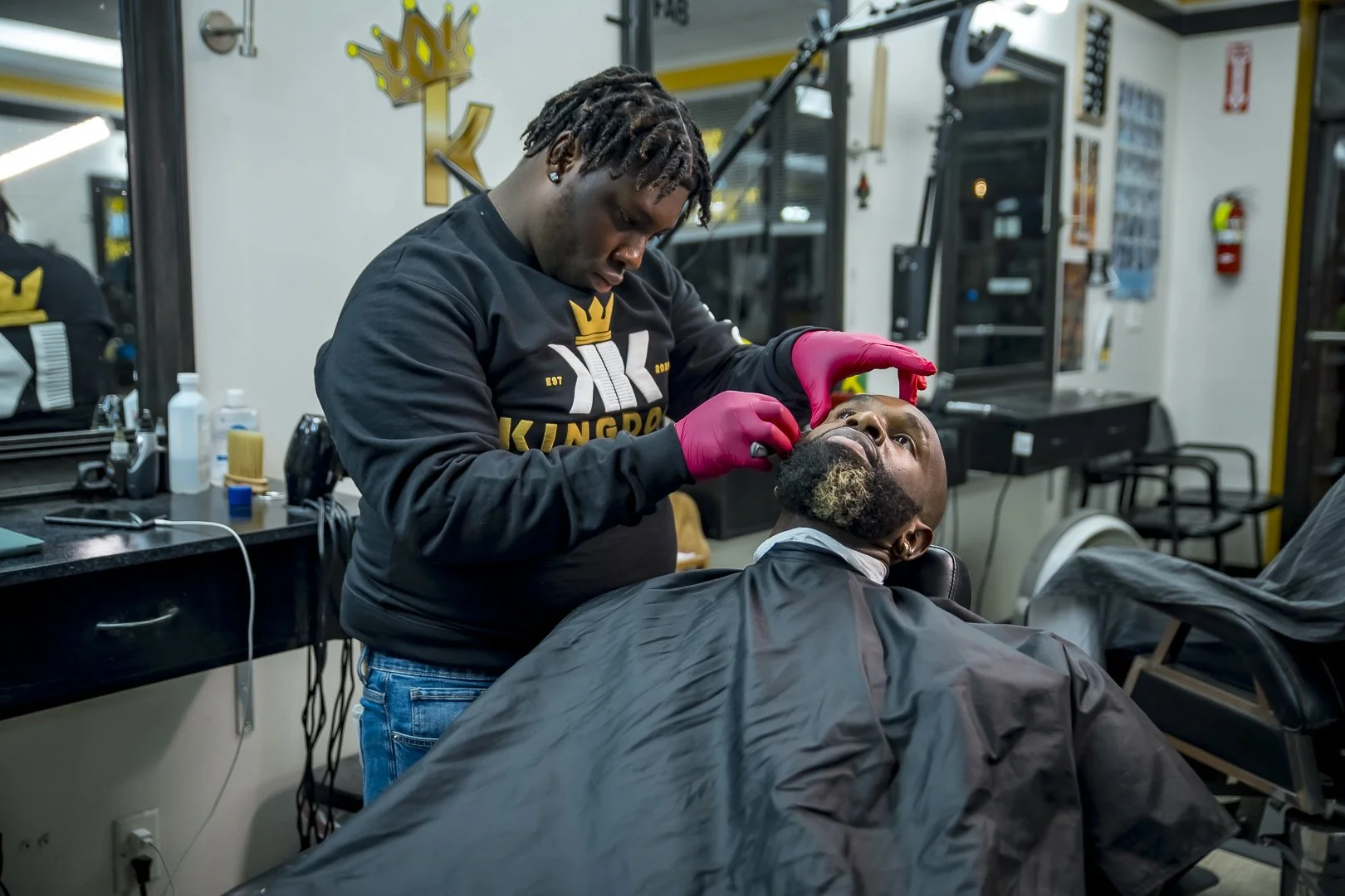 A barber wearing a black hoodie and pink gloves is trimming a man's beard in a barber shop. The man is seated in a barber chair, covered with a black cape, and looking up at the barber.