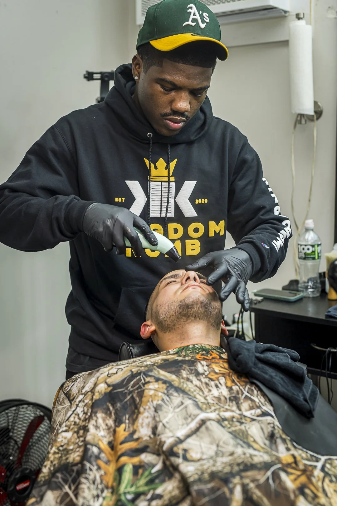 A barber wearing a black hoodie with gold and white graphics, including a crown, is giving a facial treatment to a customer in a camouflage jacket. The barber is using a handheld device near the customer's face.