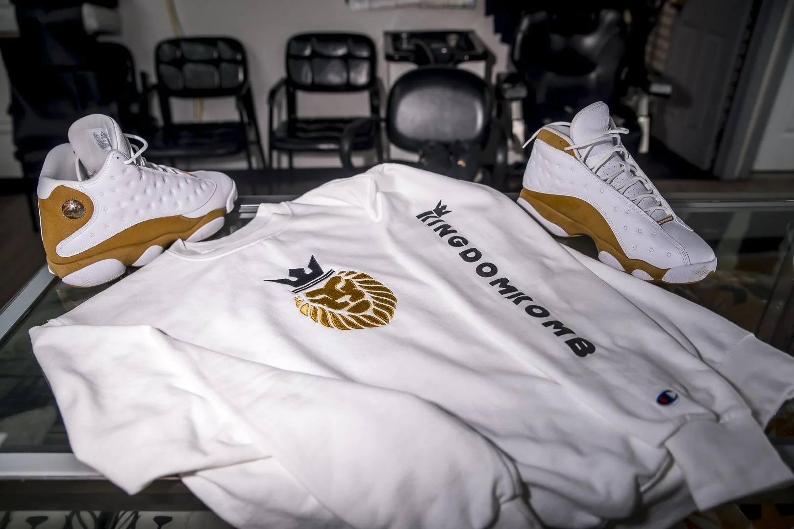 A white King Doombmb T-shirt with black logo, and matching white and brown sneakers displayed on a glass surface, with chairs in the background.