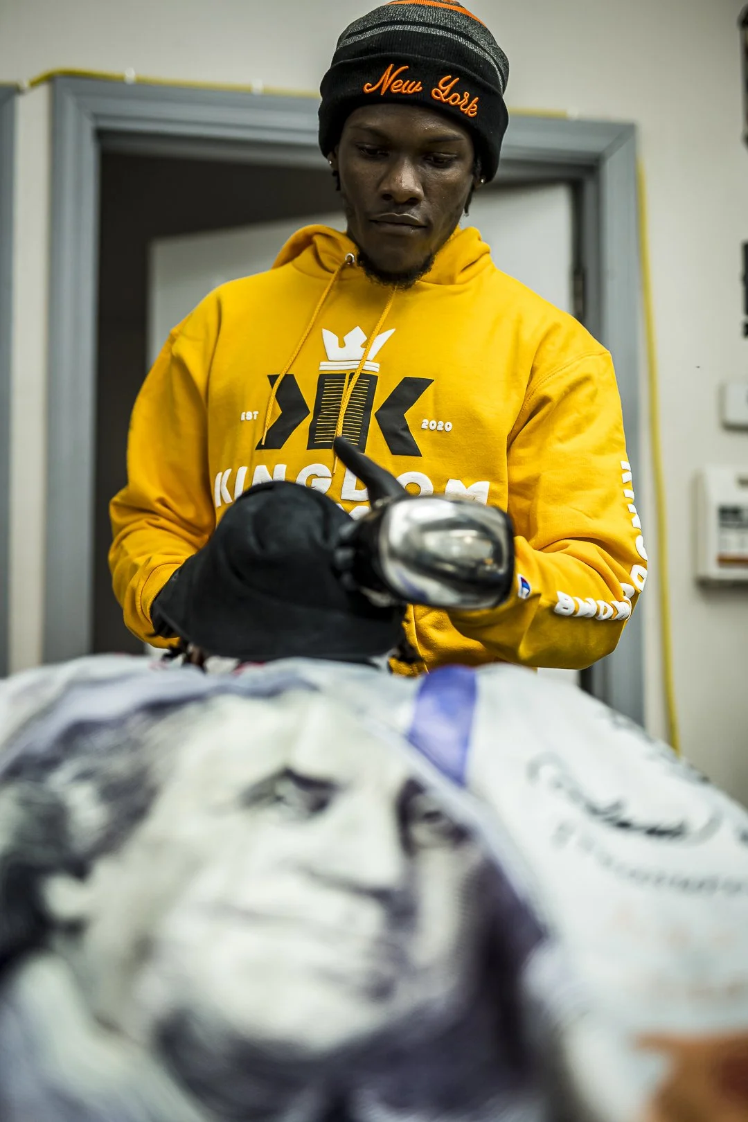 A person wearing a black beanie with orange text, a yellow hoodie, and black gloves, is looking down and working with a tool on a large item, possibly a package or bundle, in an indoor setting.