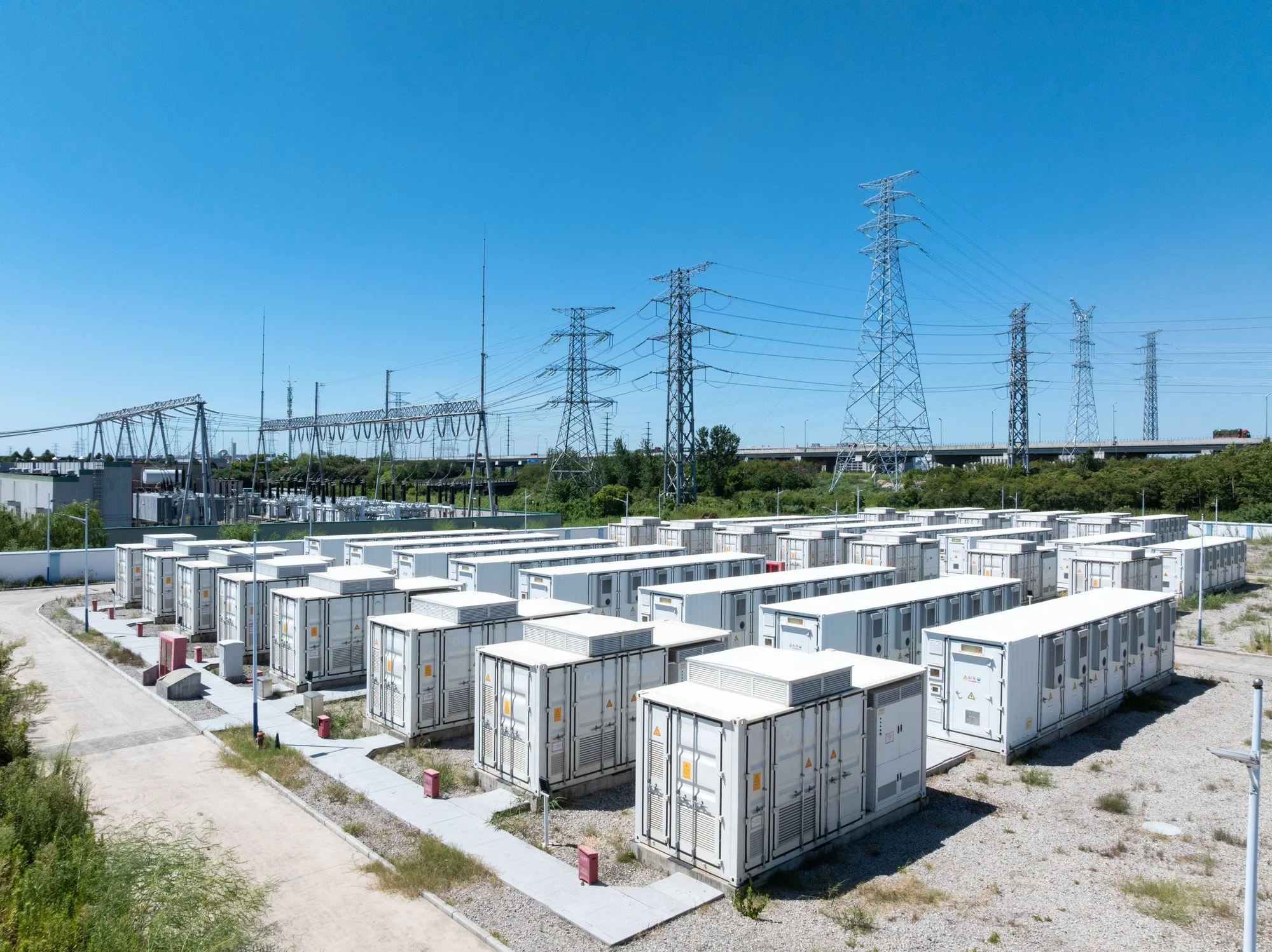 An outdoor electrical substation with multiple gray power containers, surrounded by gravel paths and small grass patches, with a backdrop of electrical towers, power lines, and a clear blue sky.