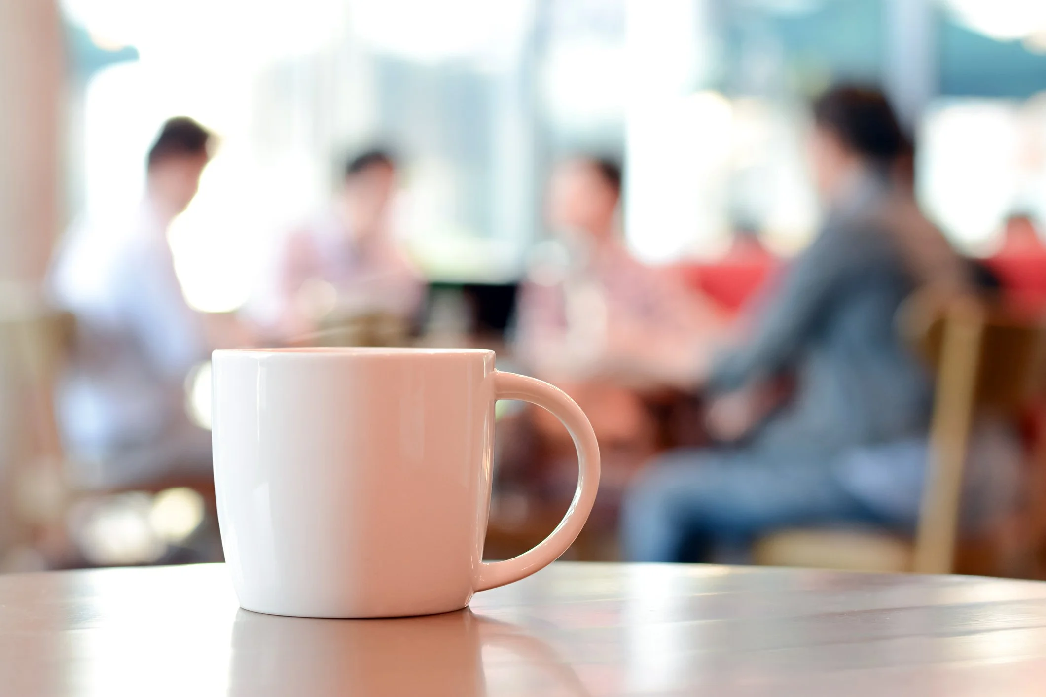 A white coffee mug on a wooden table in the foreground, with a group of four people sitting and talking at a table in a cafe or restaurant in the background, which is out of focus.