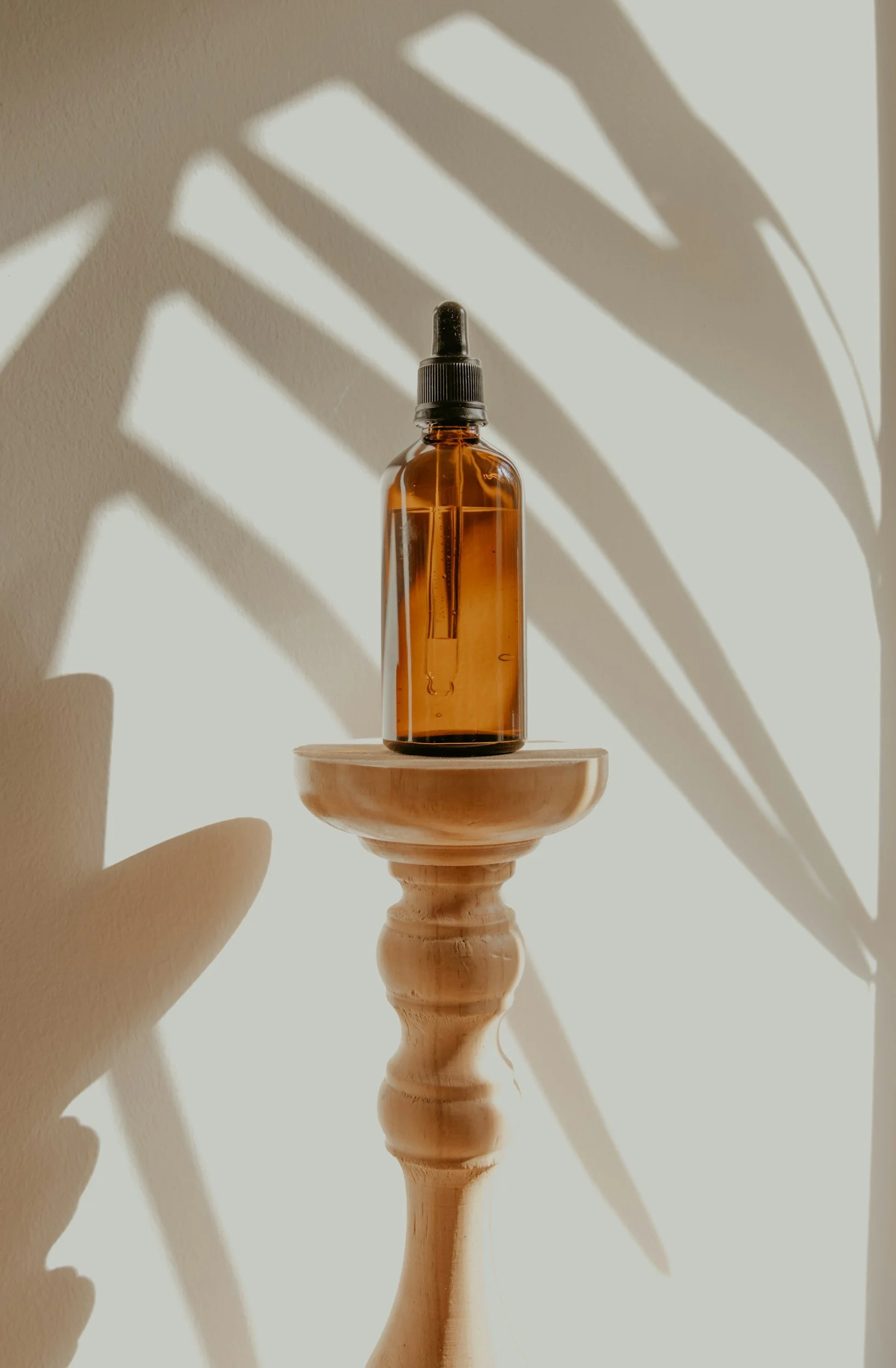 An amber glass dropper bottle on a wooden pedestal with shadow of a plant cast on the wall behind.