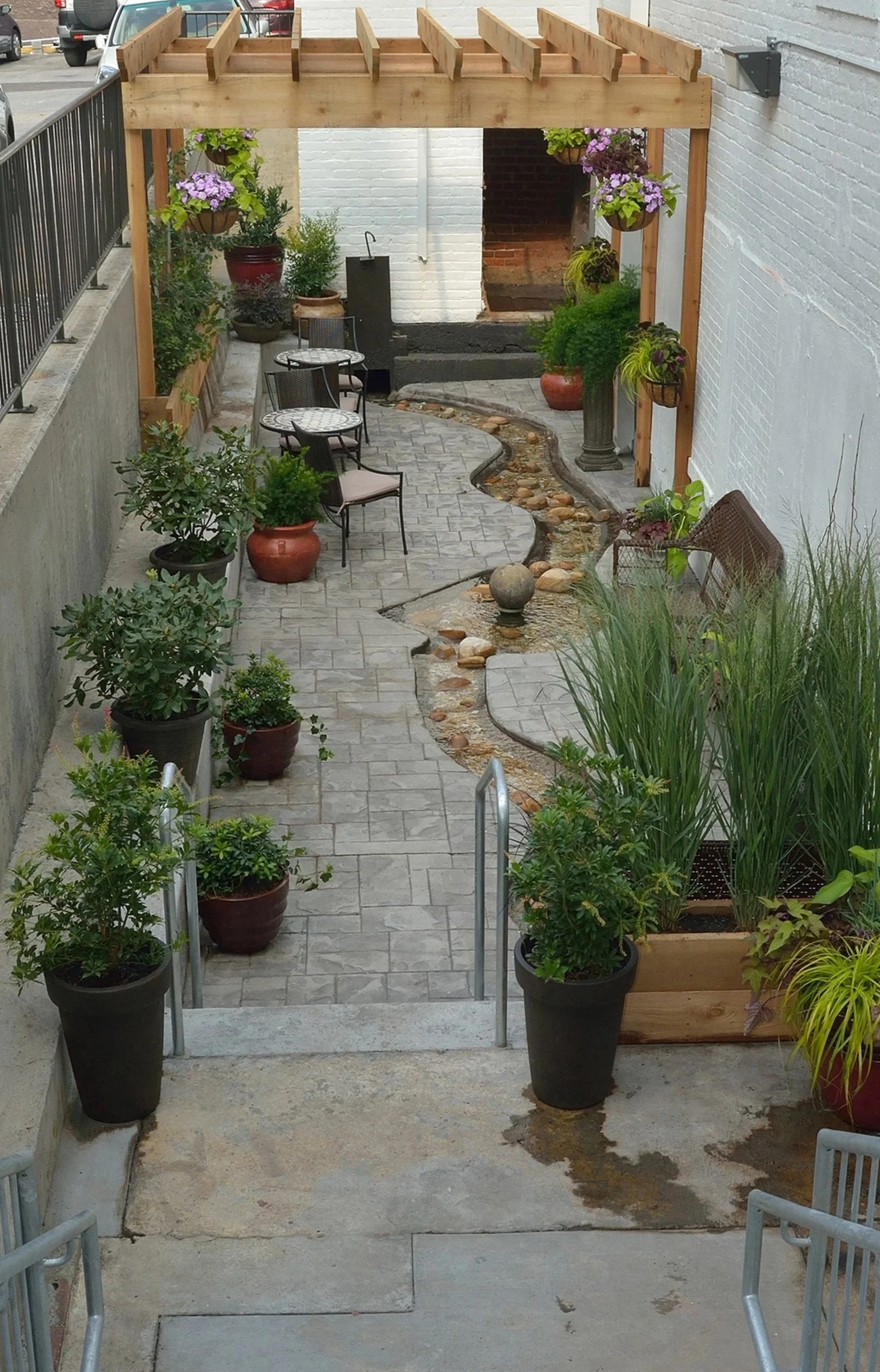 There is a paved path with potted plants, outdoor seating, a wooden pergola, and a staircase leading up to a patio area. 