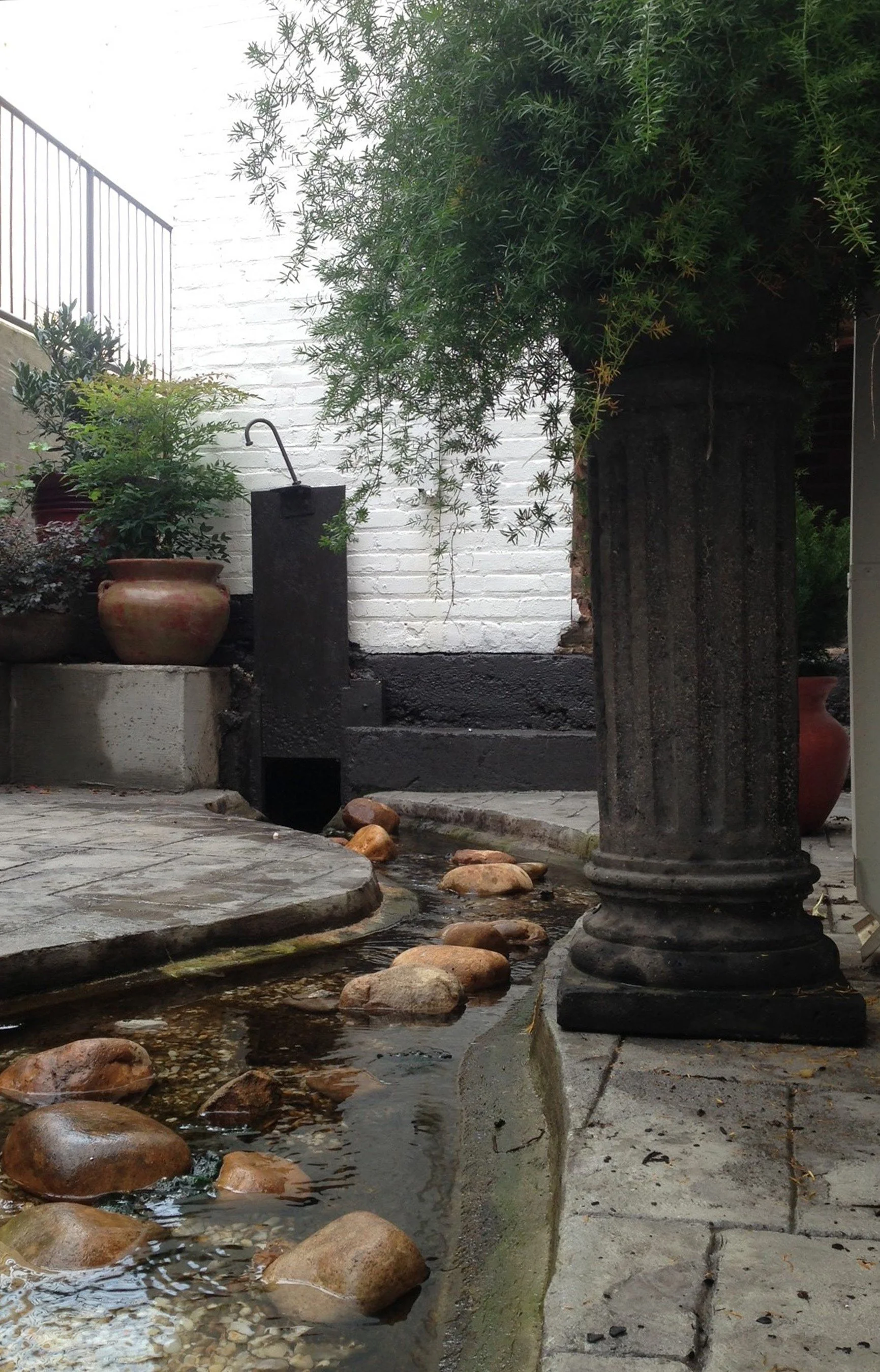 There is a small creek with rocks, a pot, a dark pedestal, and a large bush. 