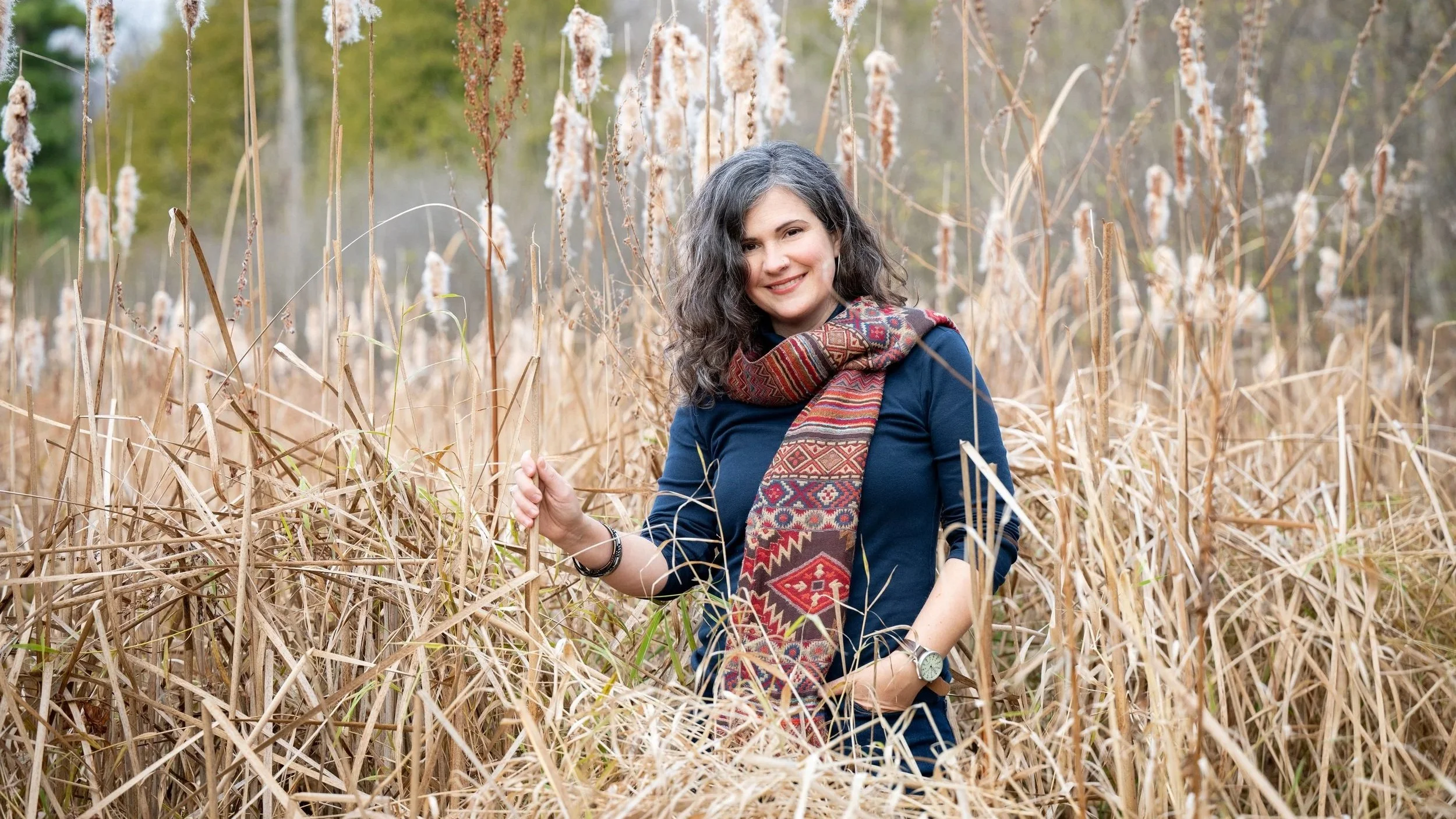 Elodie Mantha, twice-certified integrative changework coach, stands smiling in a field of tall, dry grasses, wearing a dark blue top, a colorful patterned scarf, and a watch on her left wrist.