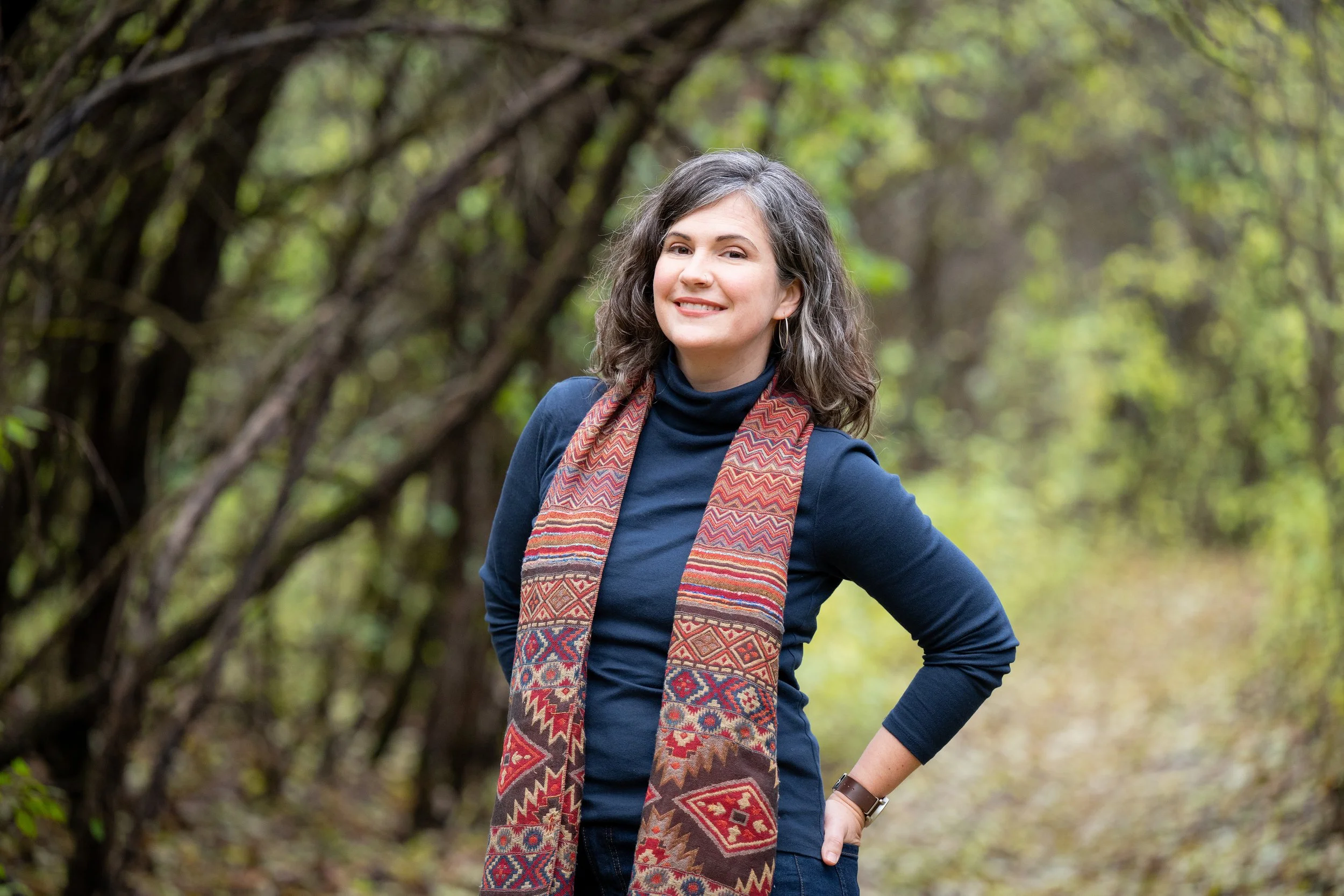 Elodie Mantha is smiling and standing on a nature trail surrounded by greenery and trees. She's wearing a navy blue turtleneck with a colorful patterned scarf, and looking at the camera directly with a confident and happy expression.