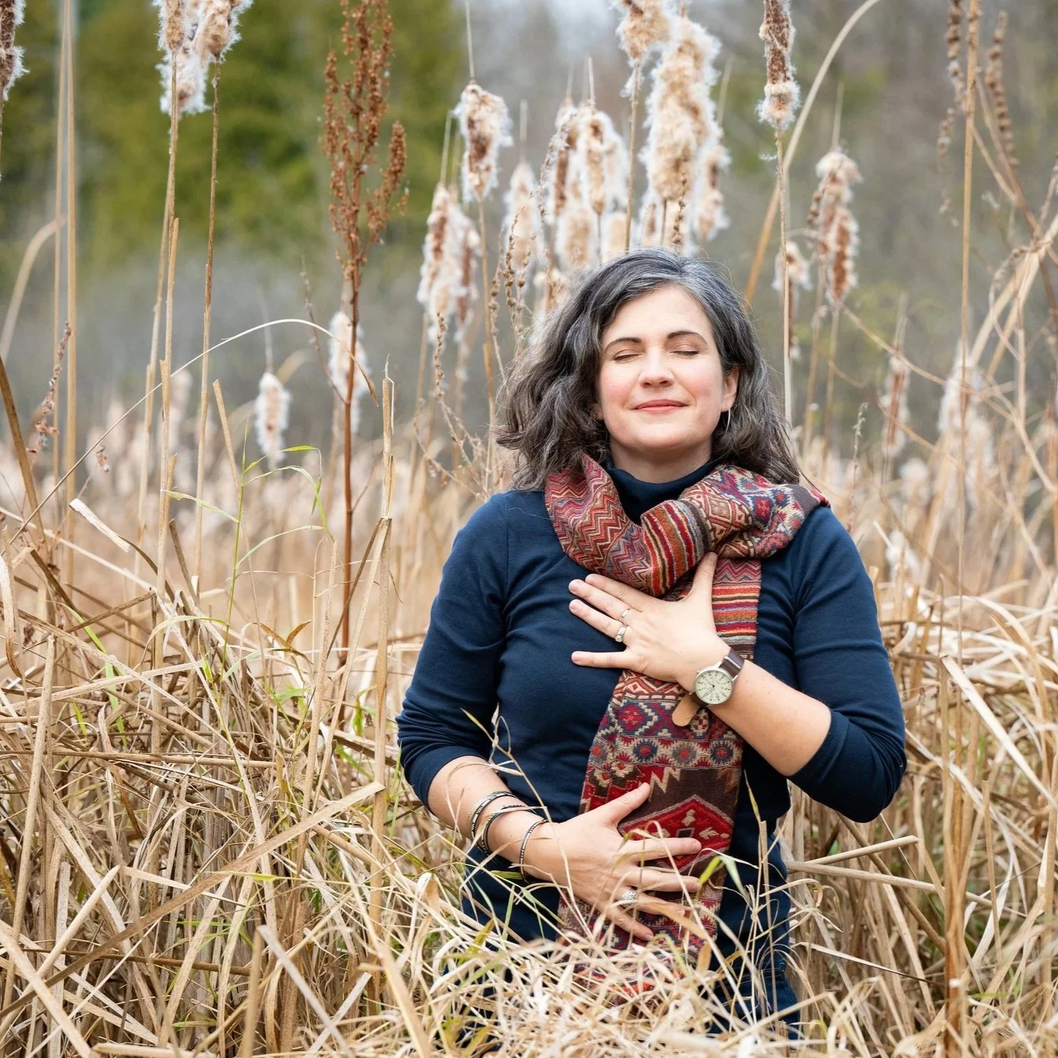 Elodie Mantha, twice-certified intergrative changework coach is standing in a field of tall, dry cattails, wearing a navy turtleneck, a red scarf and a peaceful expression, her eyes closed with one hand resting on her heart and one on her stomach.