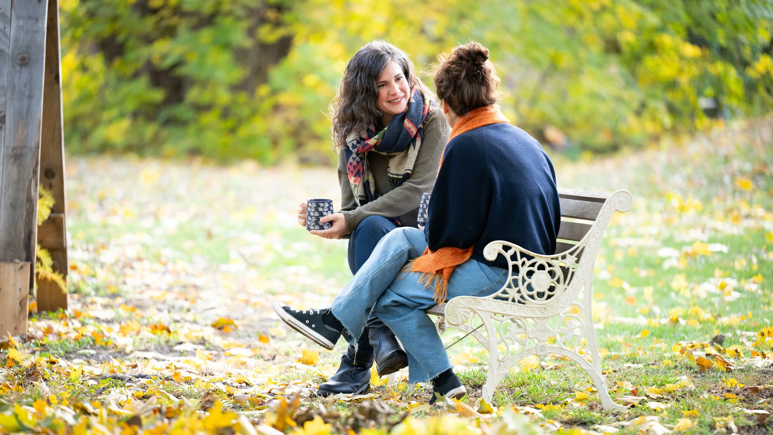 Elodie Mantha, twice-certified intergrative changework coach, sits with client on a park bench outside, engaged in a coaching conversation during autumn, surrounded by fallen yellow leaves and colorful trees.